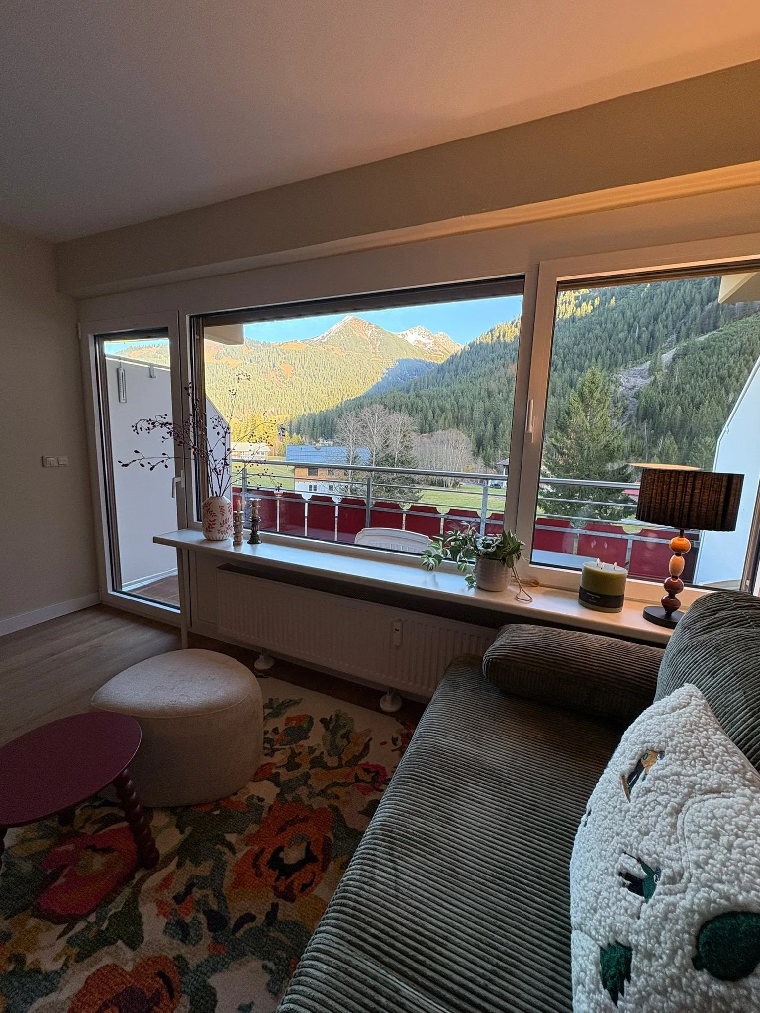 Living room with a large window showing mountain scenery, a beige ottoman, a rug with a floral pattern, a patterned sofa with a white blanket, and a side table with a lamp and plant.