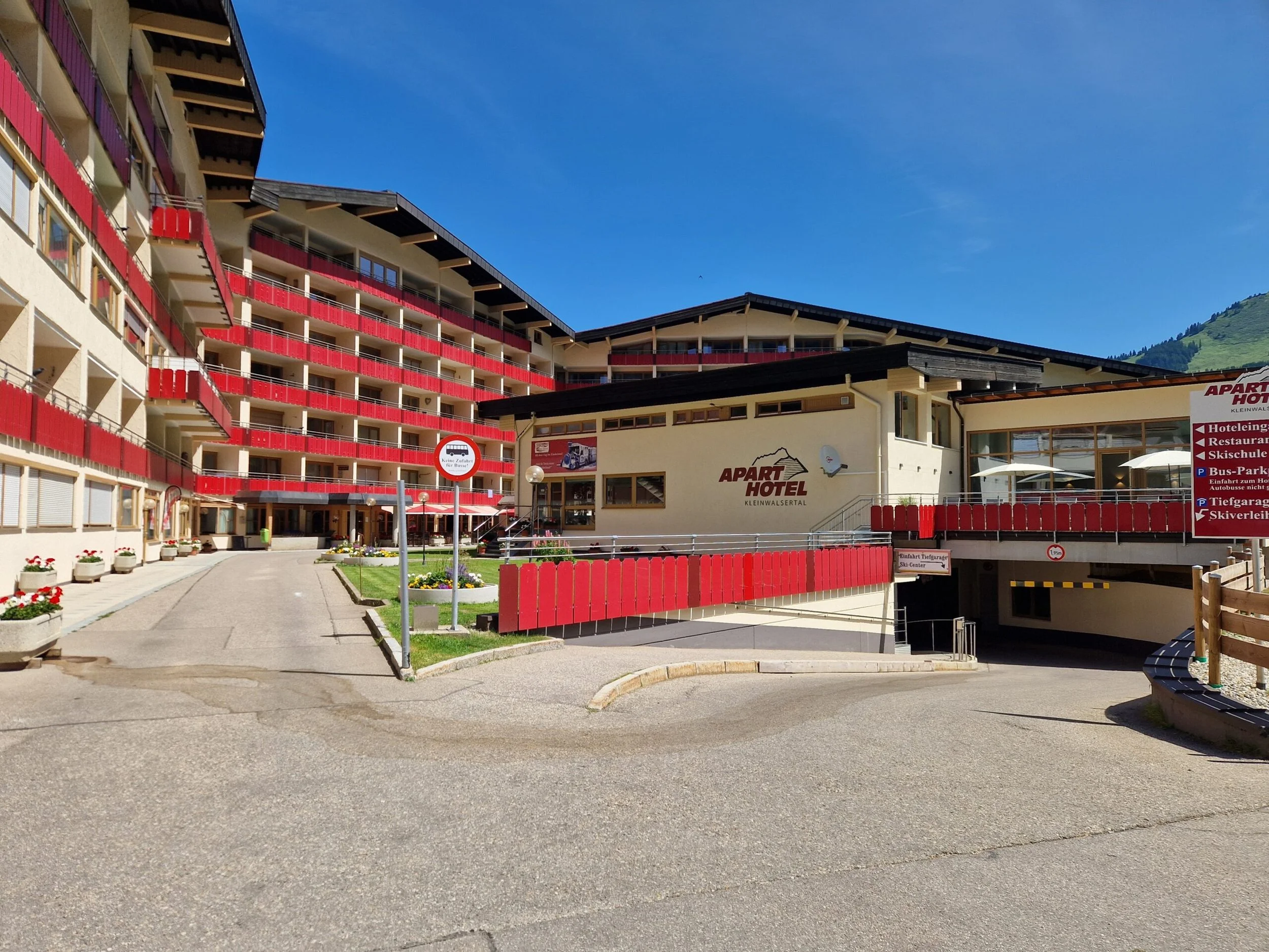 A multi-story hotel building with red balconies and a sign reading 'Apart Hotel Kleinwalsertal.' The area has a paved courtyard with flower planters and a few signs, set against a backdrop of green mountains and a clear blue sky.