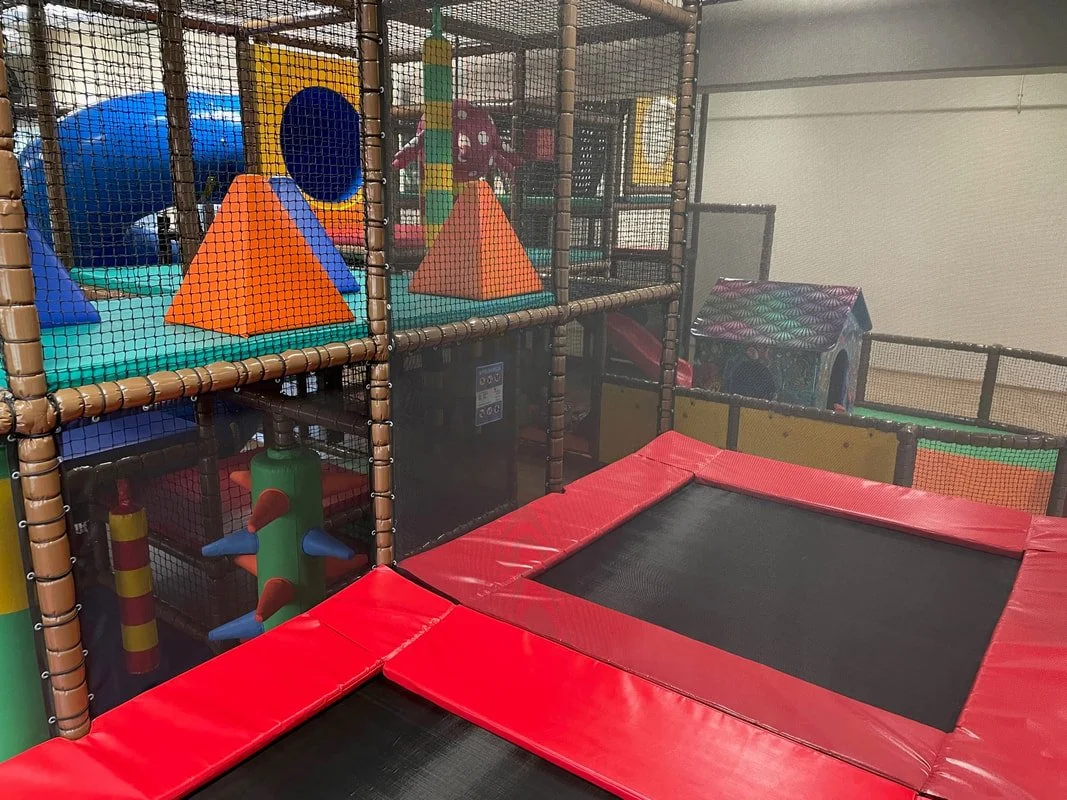 Indoor playground with colorful soft play structures, tunnels, a small house, and a trampoline with red padding.