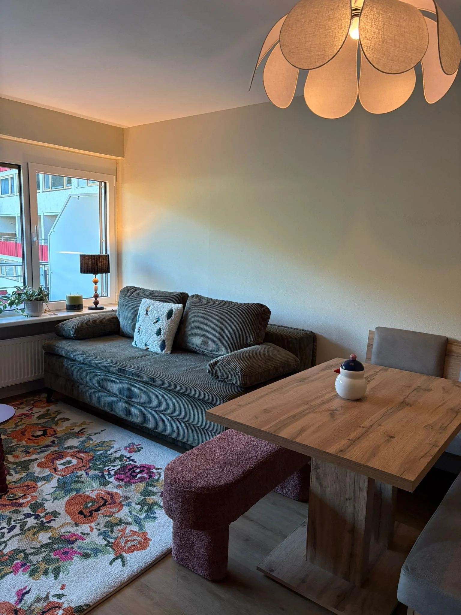 A cozy living room with a floral-patterned rug, a dark gray striped sofa adorned with white and patterned pillows, a window with plants and candles on the sill, a wooden dining table with a white ceramic teapot, a burgundy stool, a coffee table, a fl