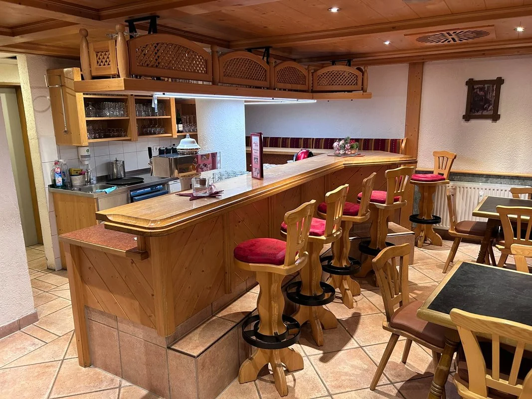 Empty restaurant with a wooden bar counter and red-cushioned bar stools. There are small tables with chairs, and a small kitchen area behind the bar with glasses, bottles, and cooking utensils. The decor includes wood paneling and a framed picture on
