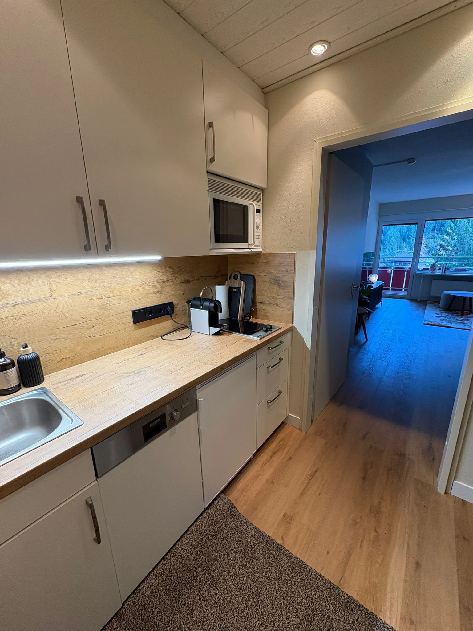 A small kitchen area with white cabinets, a wooden countertop, and a microwave oven. The kitchen opens into a living area with a balcony, which has large windows showing trees outside.