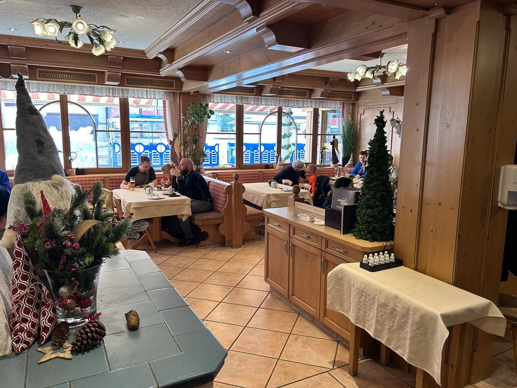 A cozy restaurant decorated for the holidays with wooden panels, holiday decorations, and a large festive holiday figure. Diners are seated at tables enjoying their meals. Outside the window, snow can be seen on the ground and bus seats with red and 