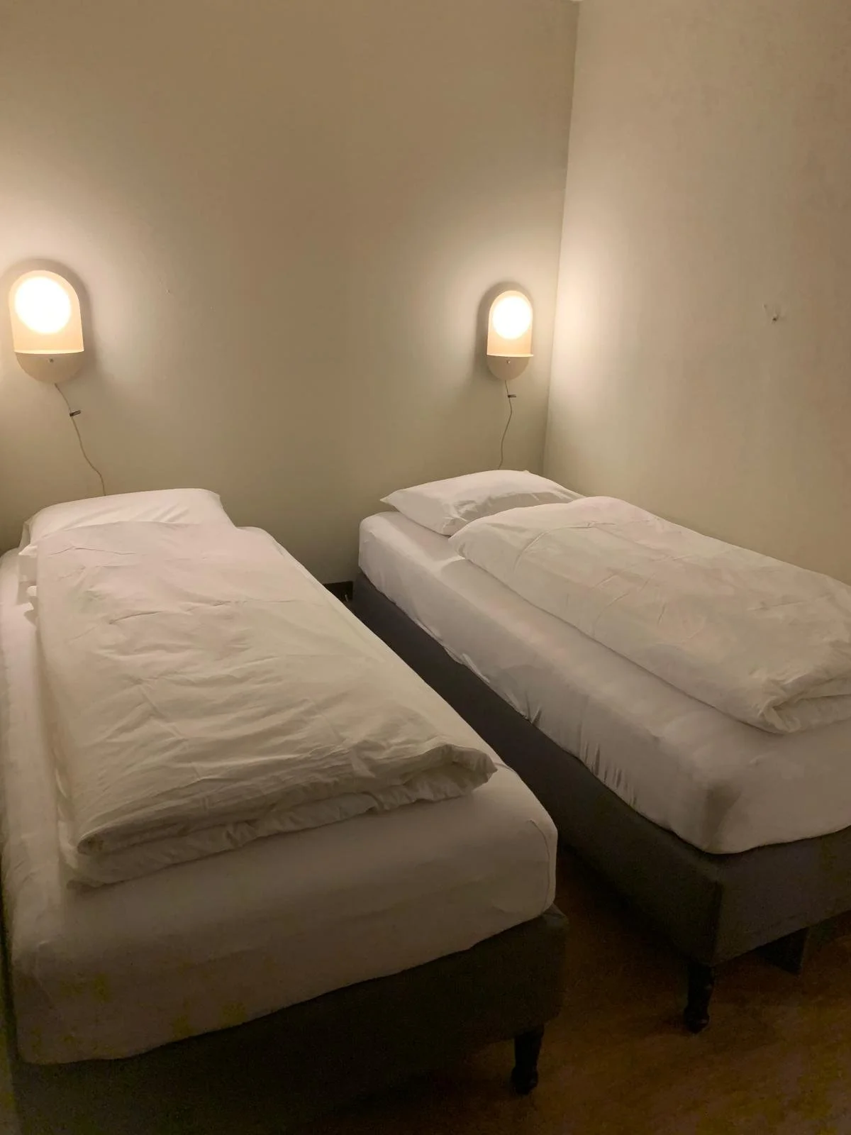 Two single beds with white sheets and pillows in a minimalist hotel room, with two wall-mounted lamps above each bed.