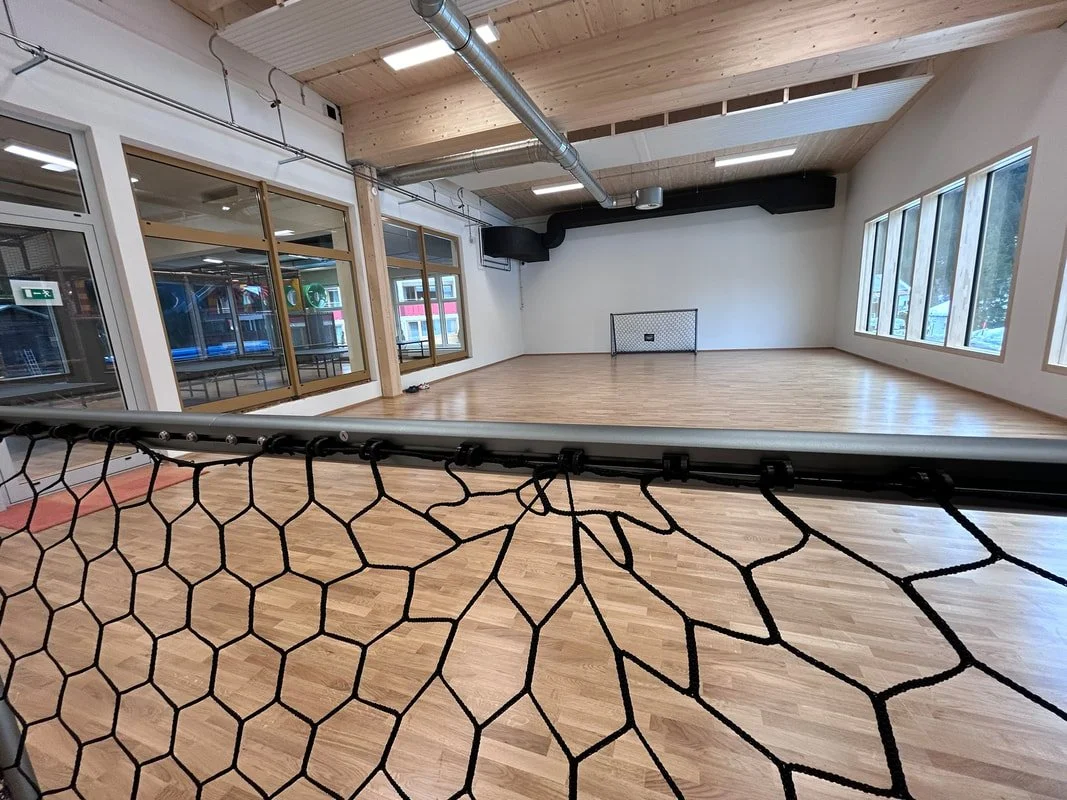 Empty indoor sports court with wooden flooring and a small hockey goal at the far end, surrounded by large windows and a netted barrier in the foreground.