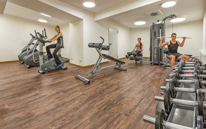 People exercising in a well-lit gym with cardio and weightlifting equipment on wood flooring.