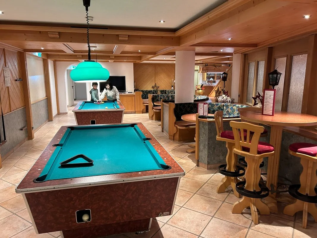 Indoor recreational area with two pool tables, a woman and two children playing pool, barstools, a round high table, seating booths, and warm wooden interior decor.