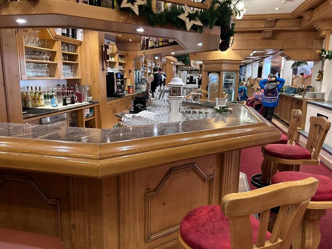 Interior of a cozy bar area decorated for Christmas with greenery and ornaments, featuring a wooden counter, red cushioned chairs, and a few people in the background.