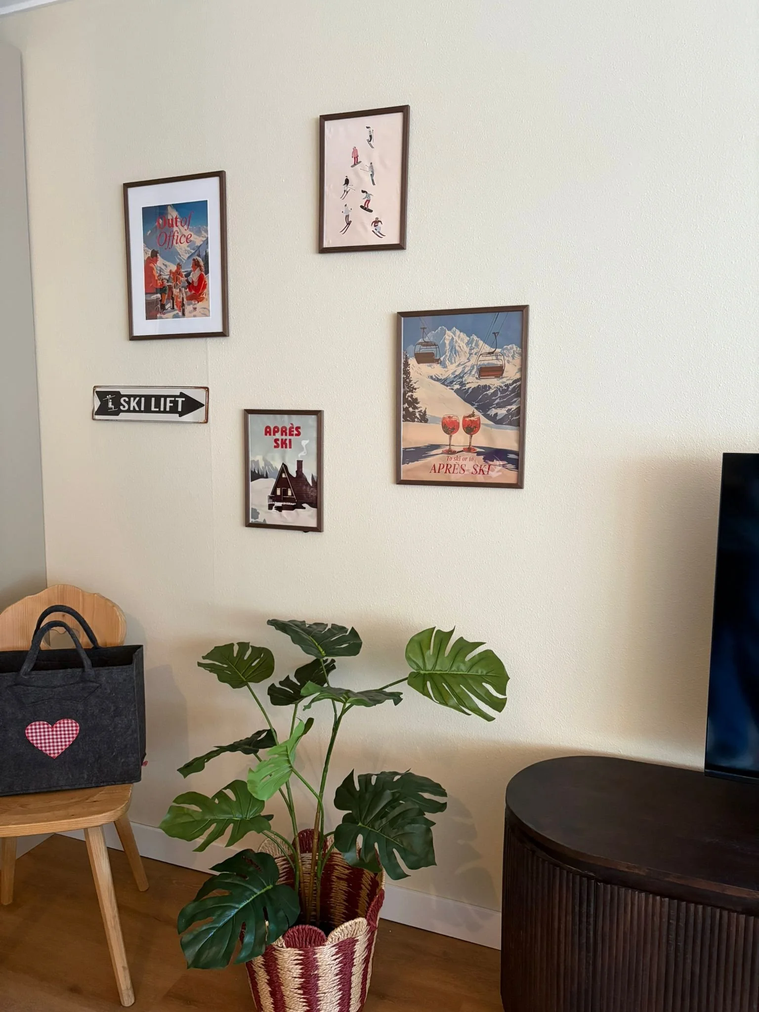 Interior of a room with framed posters on the wall, a potted plant in a basket, a wooden chair with a black paper bag, and a dark curved cabinet.