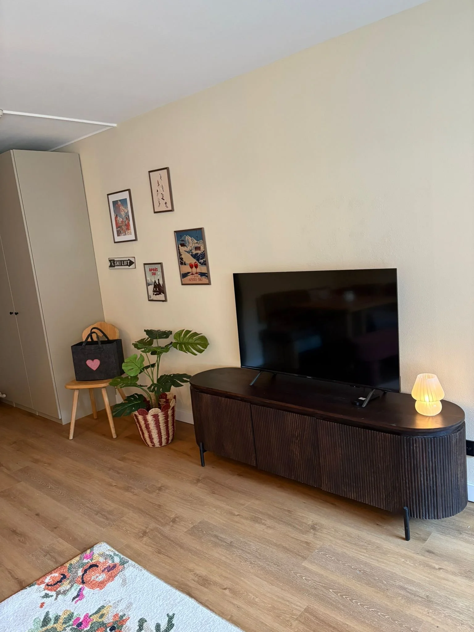 Living room with a wooden floor, a large flat-screen TV on a vintage-style wooden cabinet, a small lamp on the right, a potted plant, a stool with a black bag with a pink heart, and framed art on the beige wall.