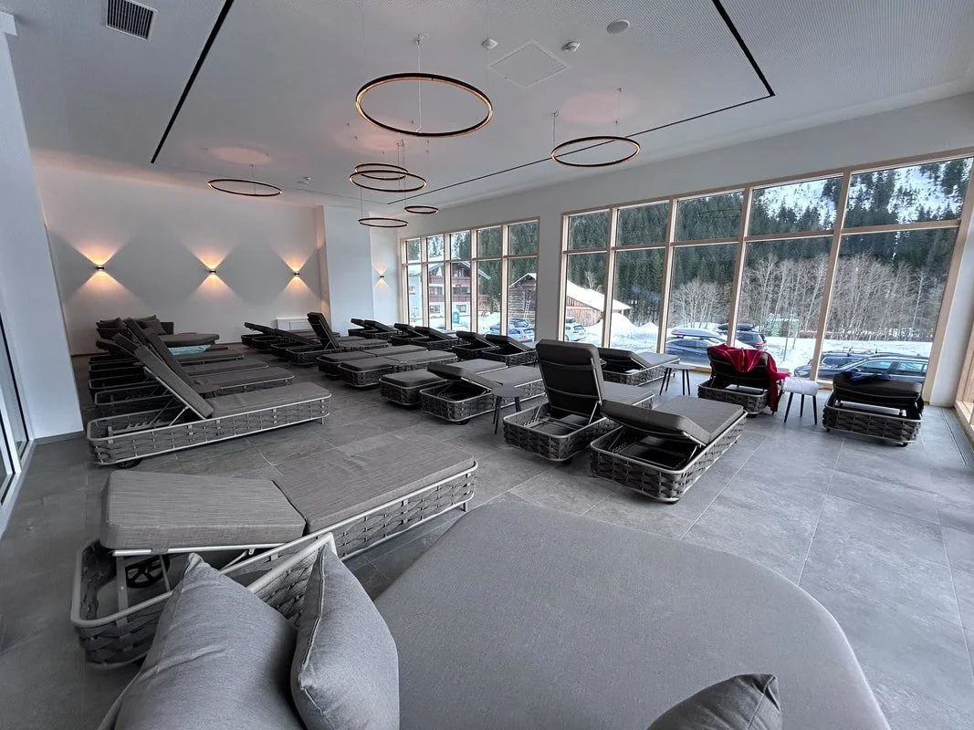 Modern lounge with lounge chairs and tables, large windows showing snowy landscape outside, decorative ceiling lights, and soft wall lighting.