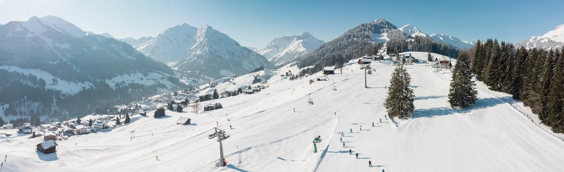 Snow-covered ski resort with ski lifts, skiers, pine trees, and wooden buildings, set against a backdrop of snow-capped mountains.