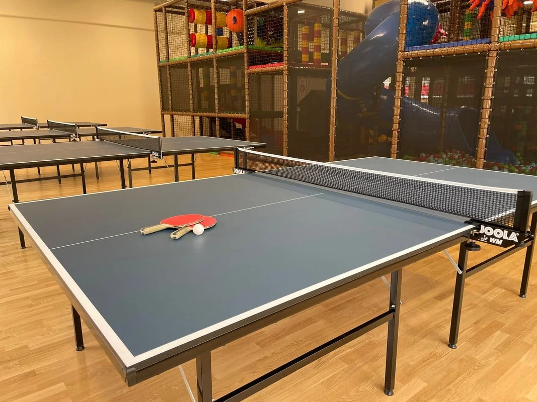 Indoor room with multiple ping pong tables set up for play, with paddles and balls on the closest table, and a colorful indoor playground structure in the background.