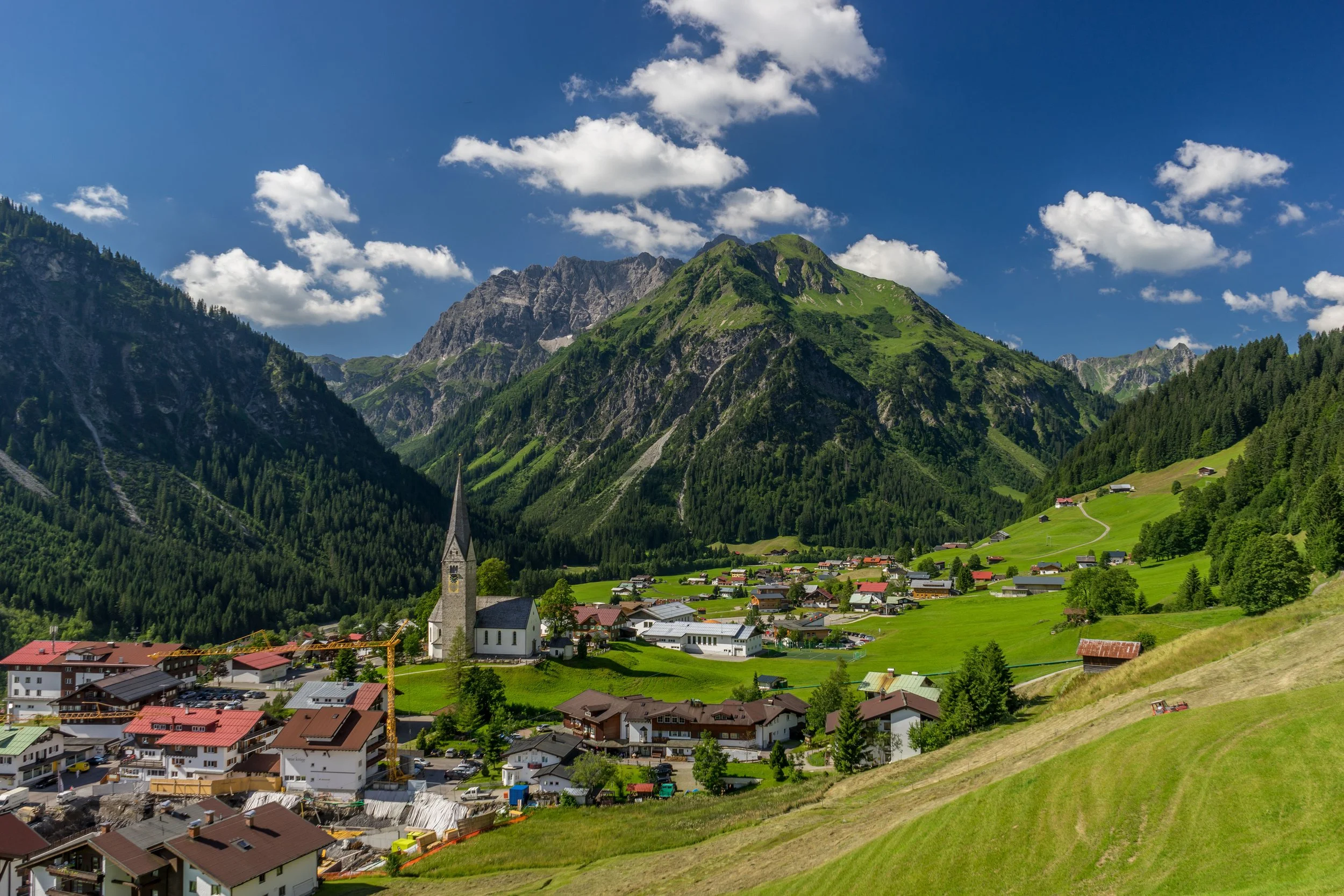 Scenic mountain village with green hills, colorful houses, a church with a tall steeple, and towering mountains in the background under a blue sky with white clouds.