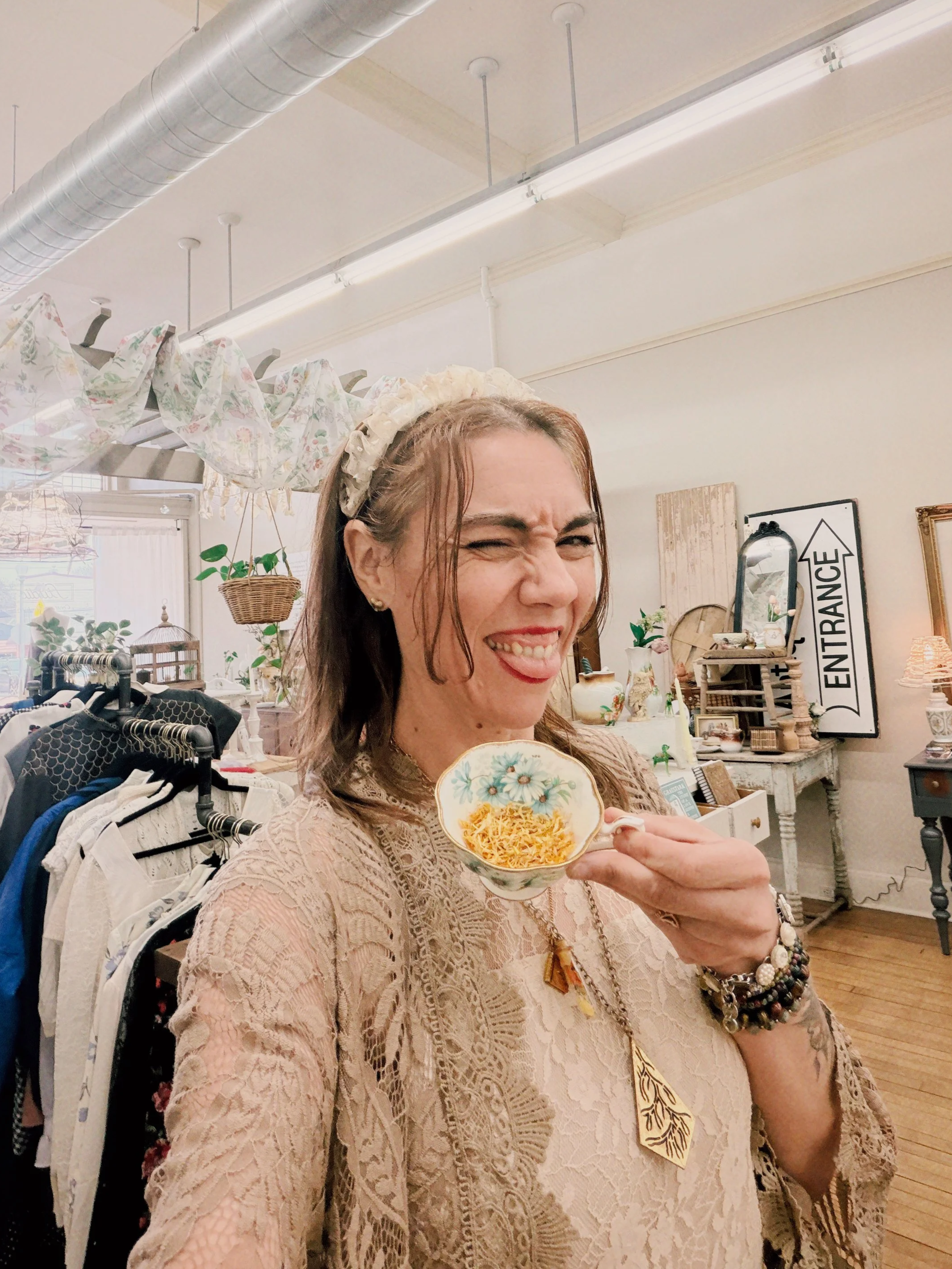 A woman wearing a lace beige top and flower headband is holding a floral patterned bowl with shredded cheese, smiling and making a face in a boutique with clothing racks and decorative items in the background.