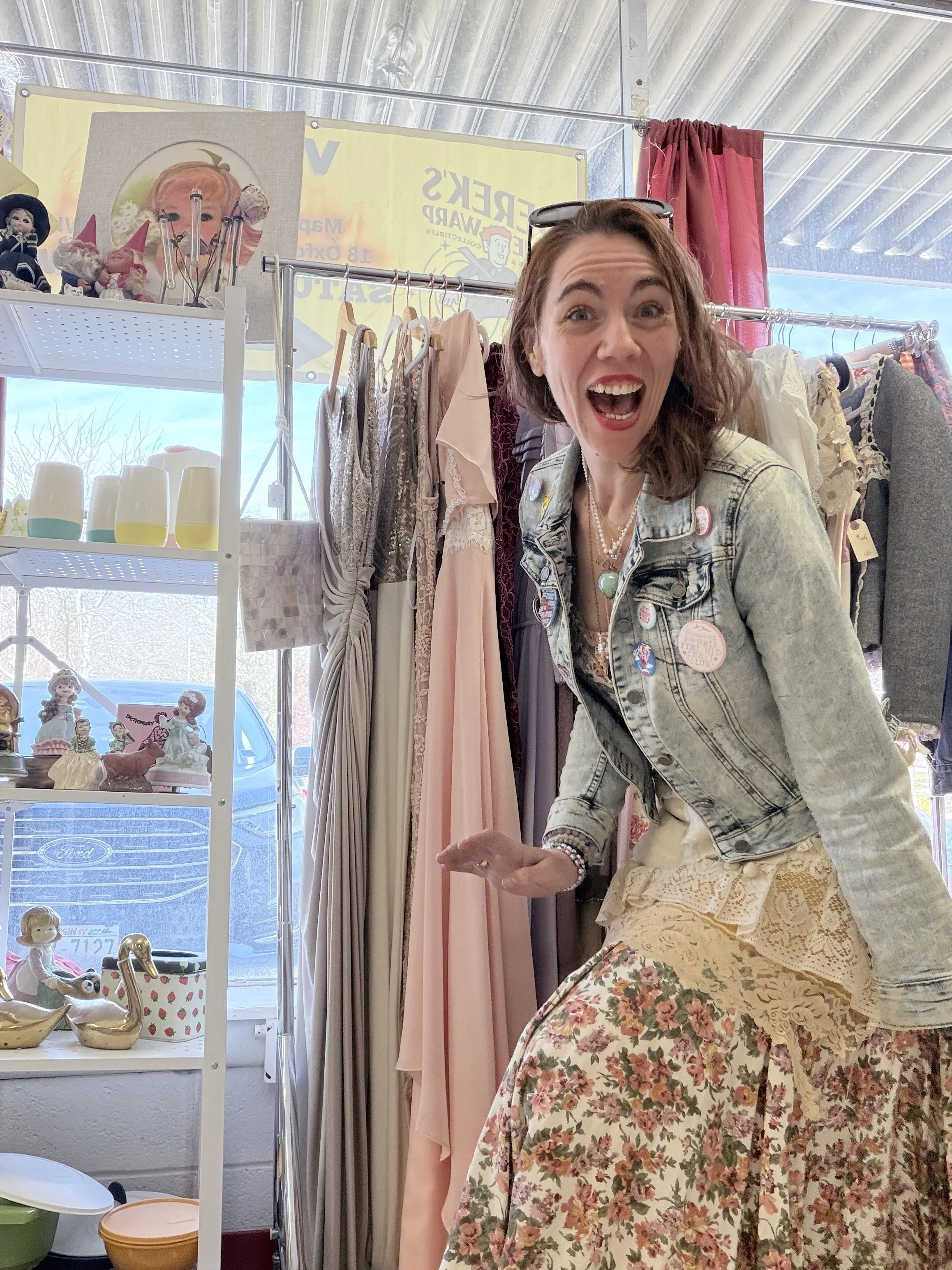A woman with long brown hair smiling and holding a phone inside a boutique store with clothes on racks and decorative items on shelves.