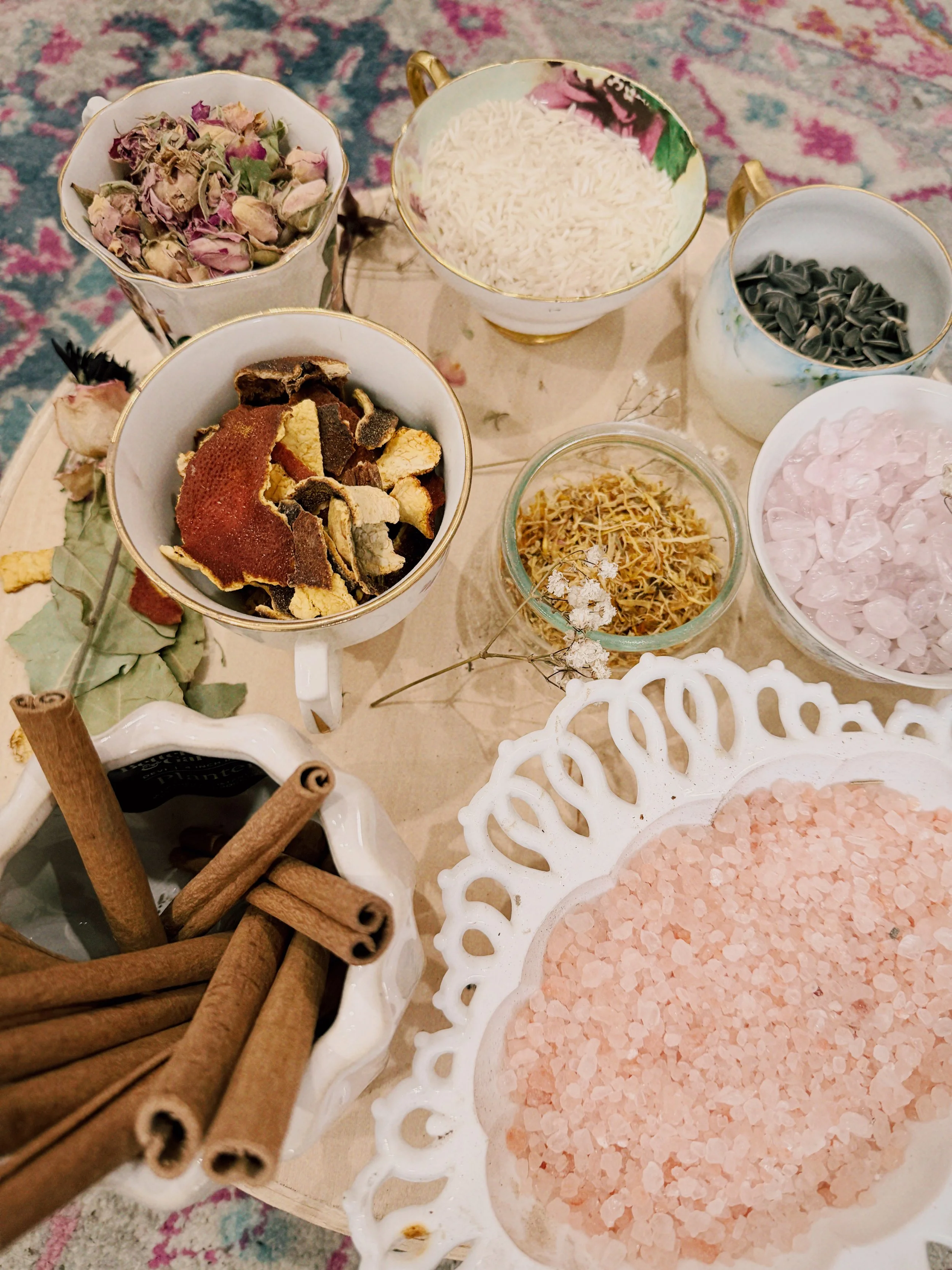 A collection of bowls filled with dried flowers, rice, sunflower seeds, crystal rocks, and pink Himalayan salt, arranged on a table with a white decorative tray and cinnamon sticks.