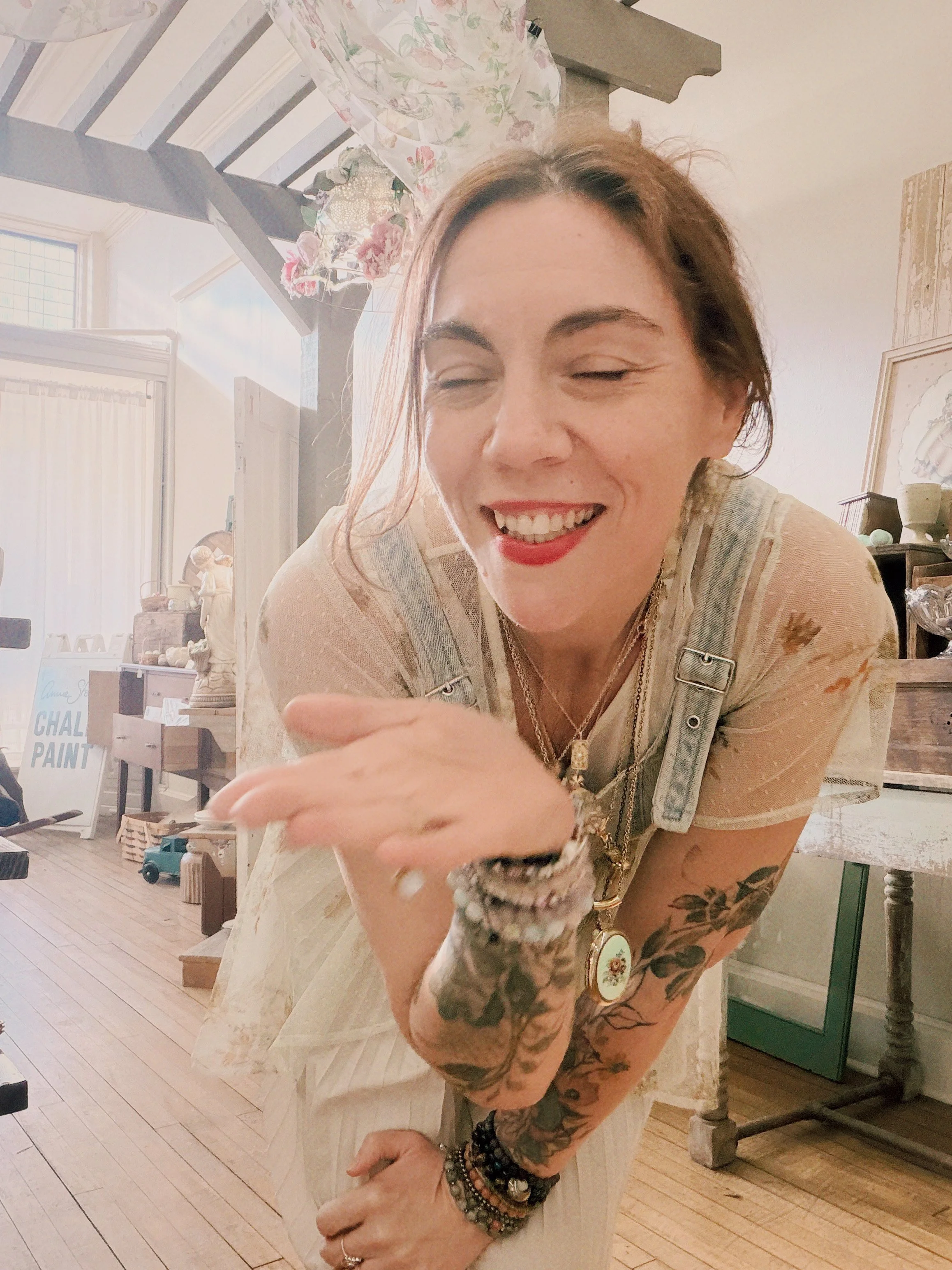 A woman with tattoos on her arms and wearing multiple bracelets, a necklace, and a ring, smiling with eyes closed, in a room with white walls and wooden floors, surrounded by decorative items and furniture.