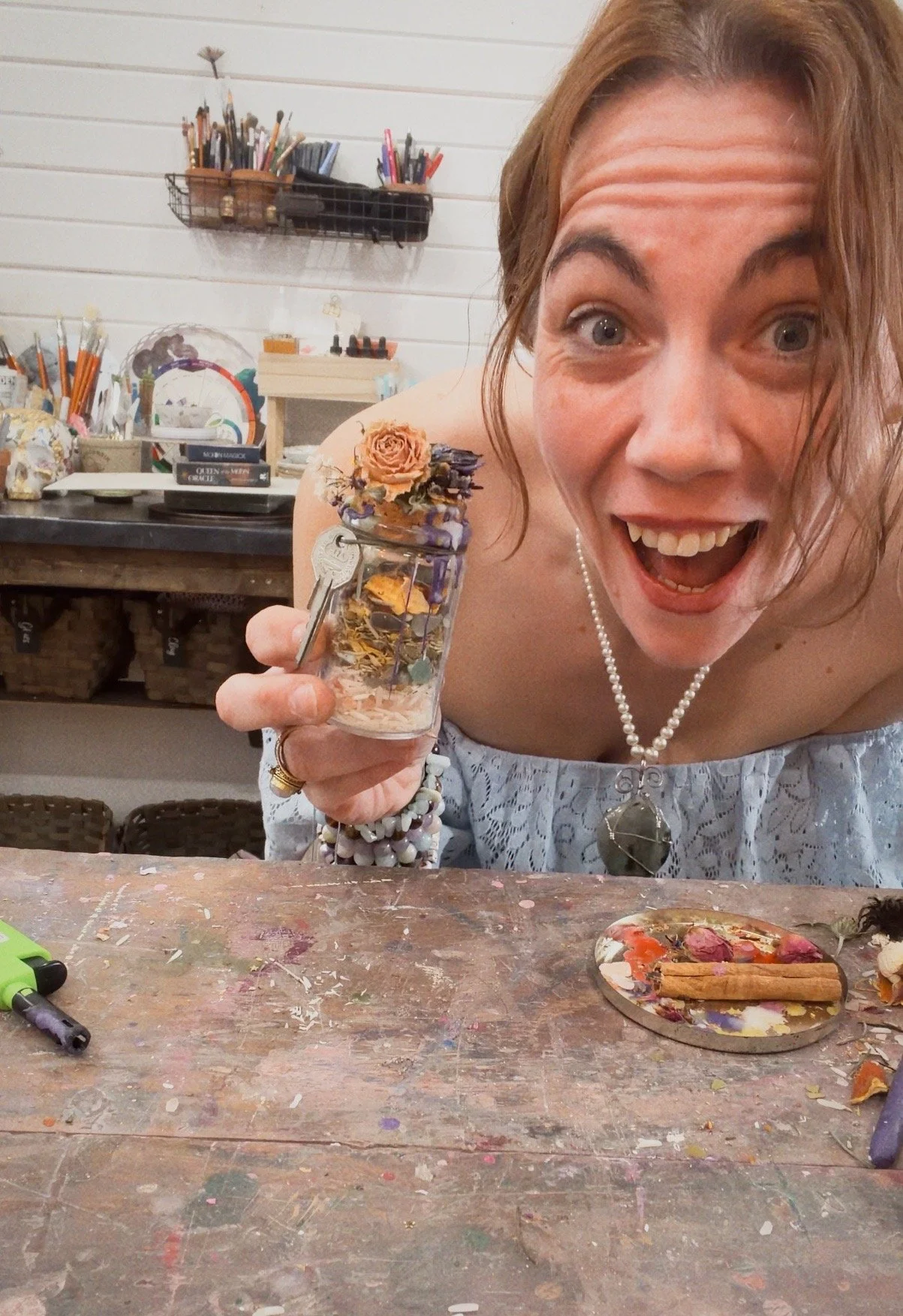A woman with excited expression holding a small jar filled with dried flowers, herbs, and decorative items, in a craft or art studio with art supplies and brushes in the background.
