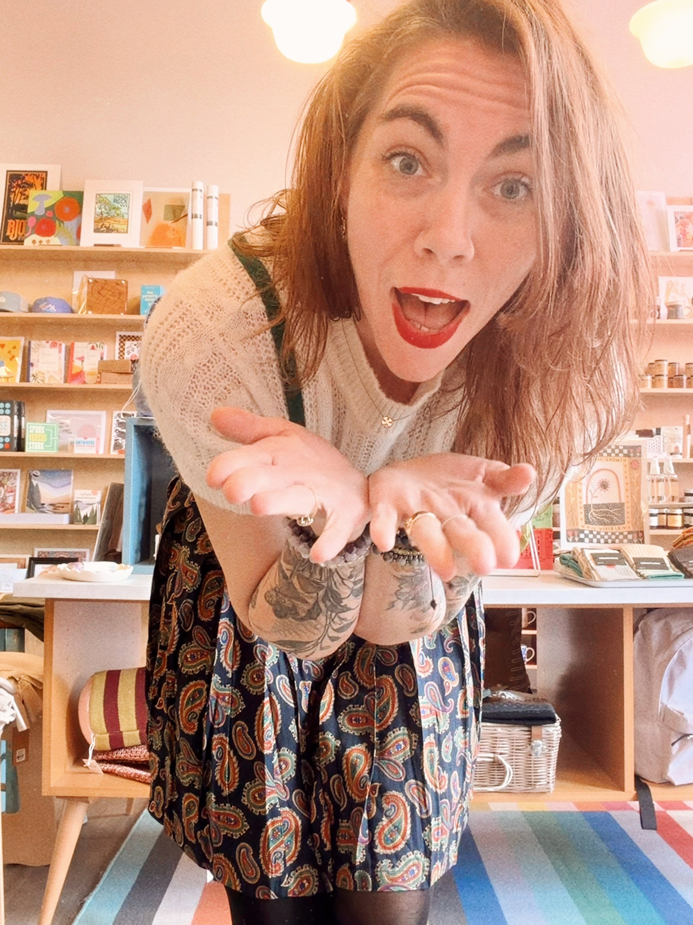 A woman with red hair and tattoos, wearing a white sweater, patterned skirt, and red lipstick, leaning forward with an excited expression in a colorful store or cafe with wood shelves and various items.