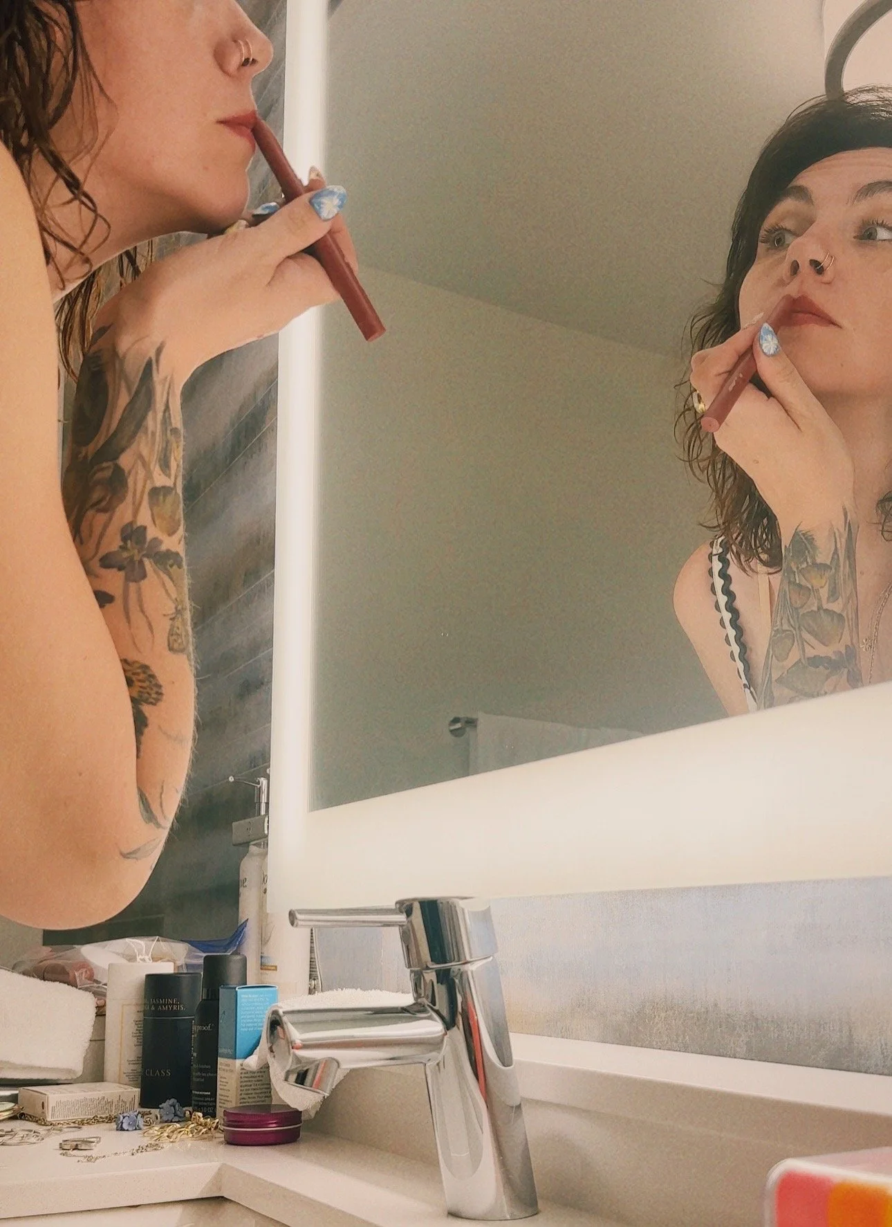 A woman with tattoos applying lip balm while looking at herself in a bathroom mirror. There are various toiletries on the bathroom counter including bottles, jewelry, and a towel.