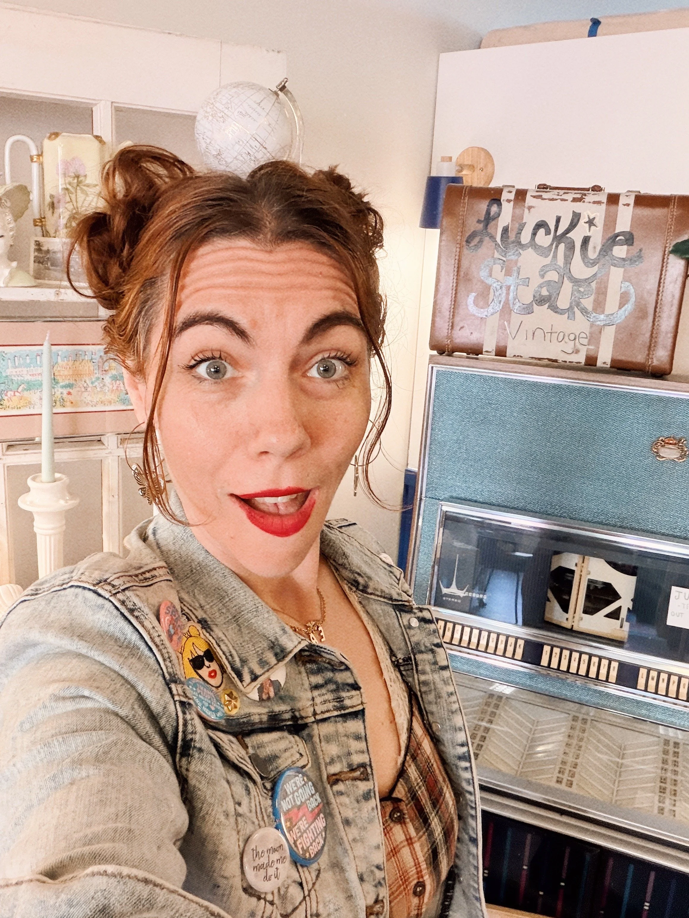 Young woman with red hair wearing a denim jacket and multiple buttons, taking a selfie in front of vintage items, including a jukebox and a sign reading "Lucky Star Vintage."
