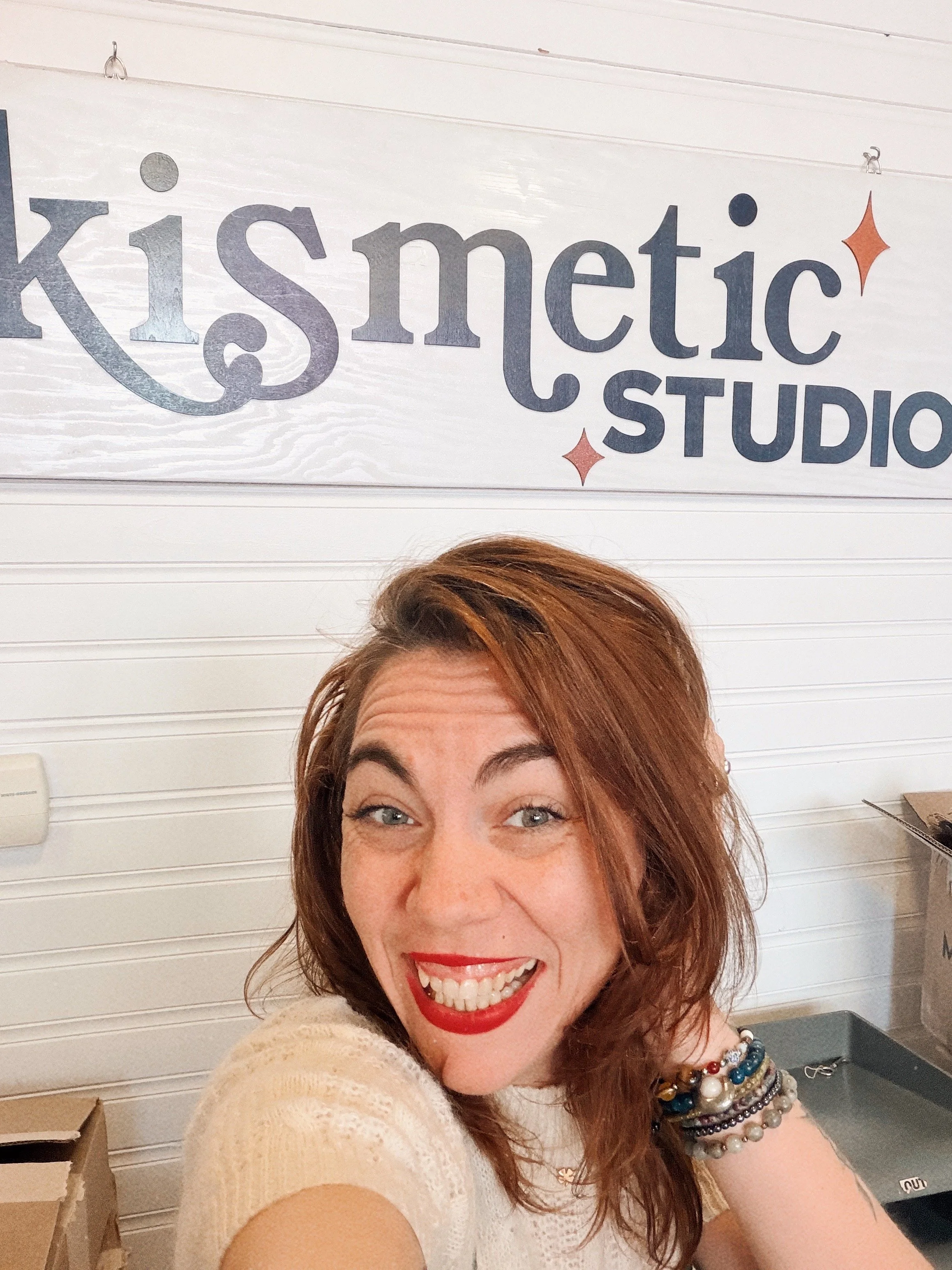Close-up of a smiling woman with red hair and multiple bracelets on her wrist, in front of a sign that says 'kismet STUDIO'.