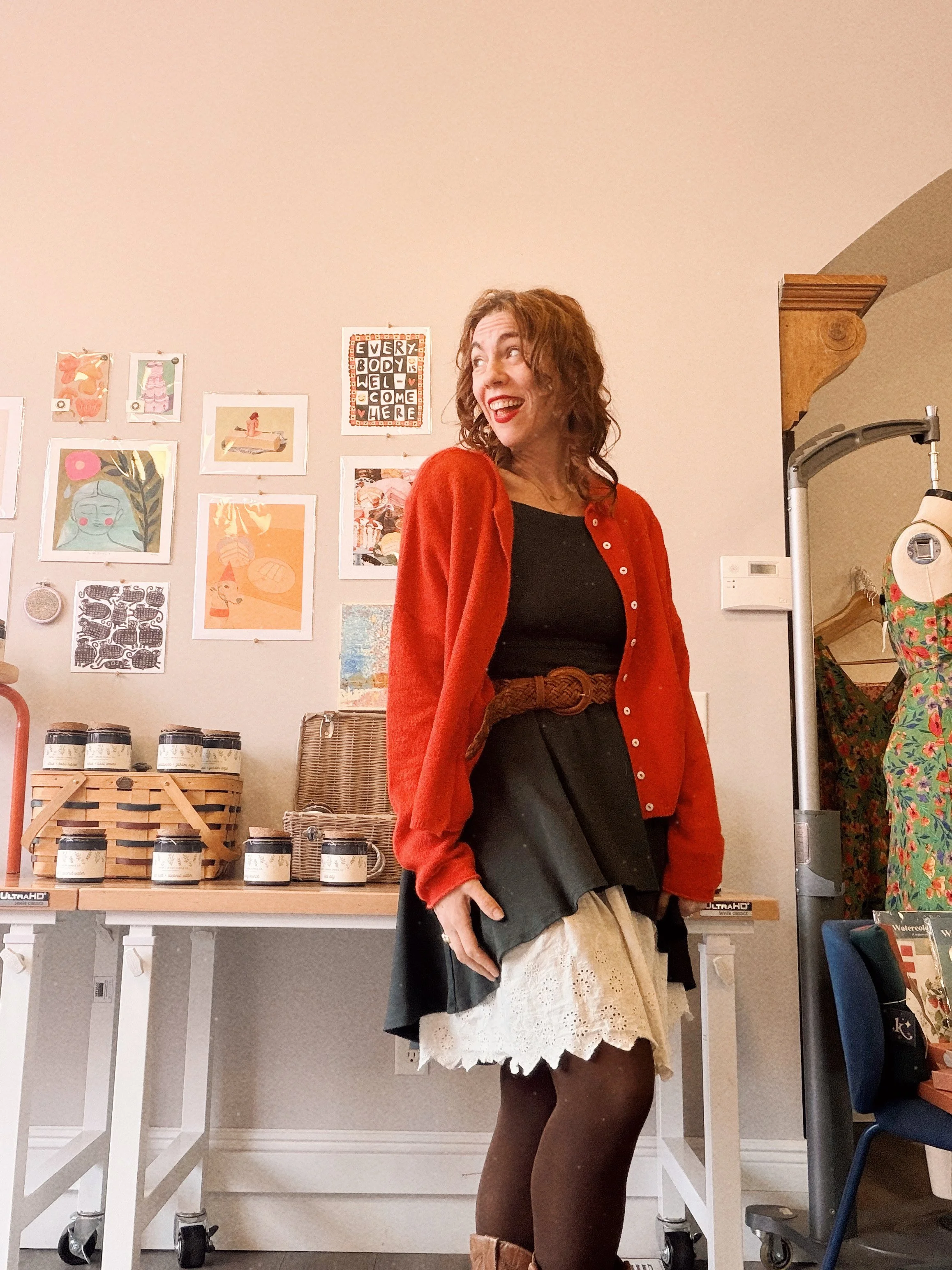 A woman with curly brown hair wearing a red cardigan and black skirt, holding a beige handbag, standing in an art gallery or store with colorful artwork on the wall behind her.