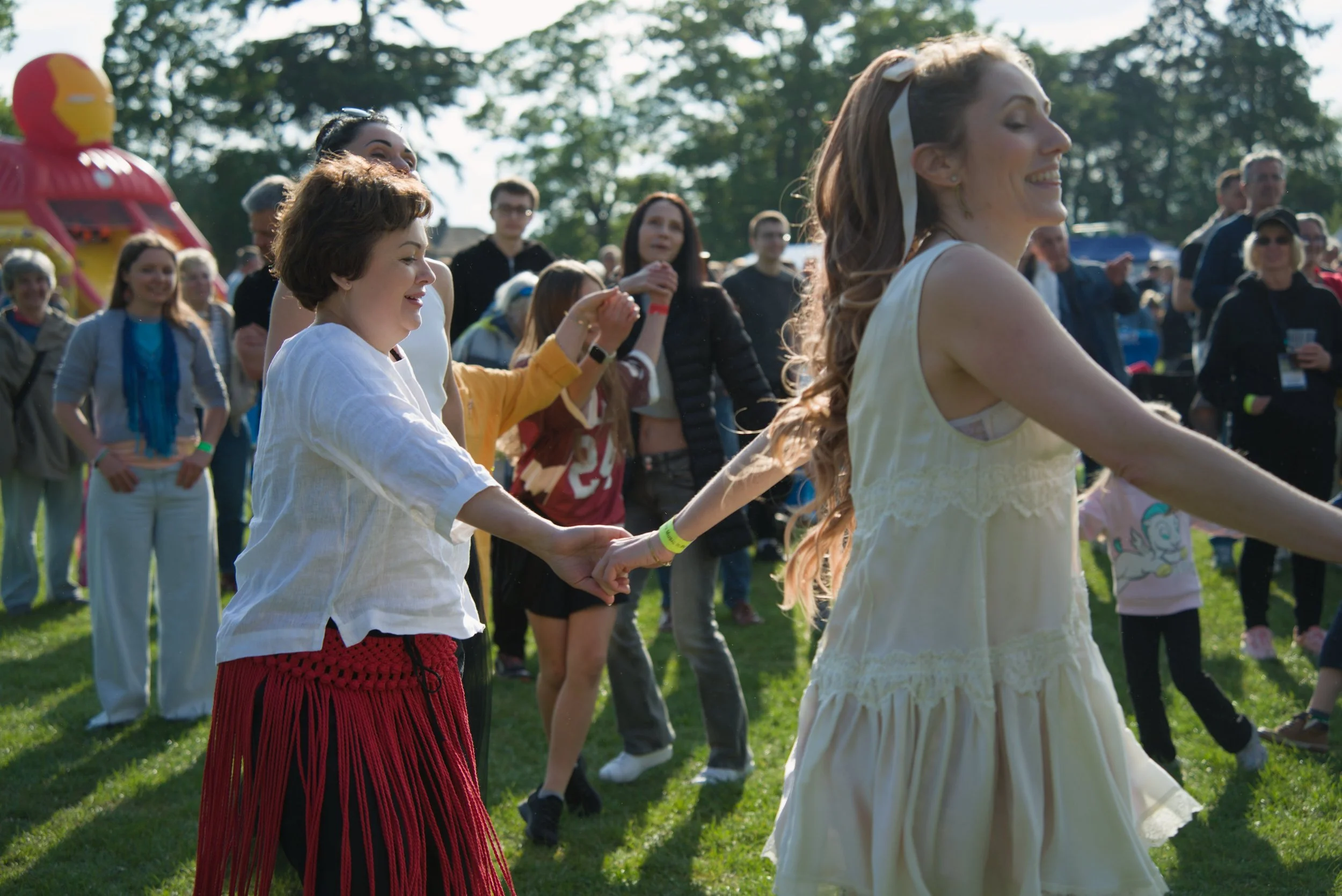 People dancing outdoors during the daytime, holding hands in a circle, at a festival or gathering, with green trees in the background.