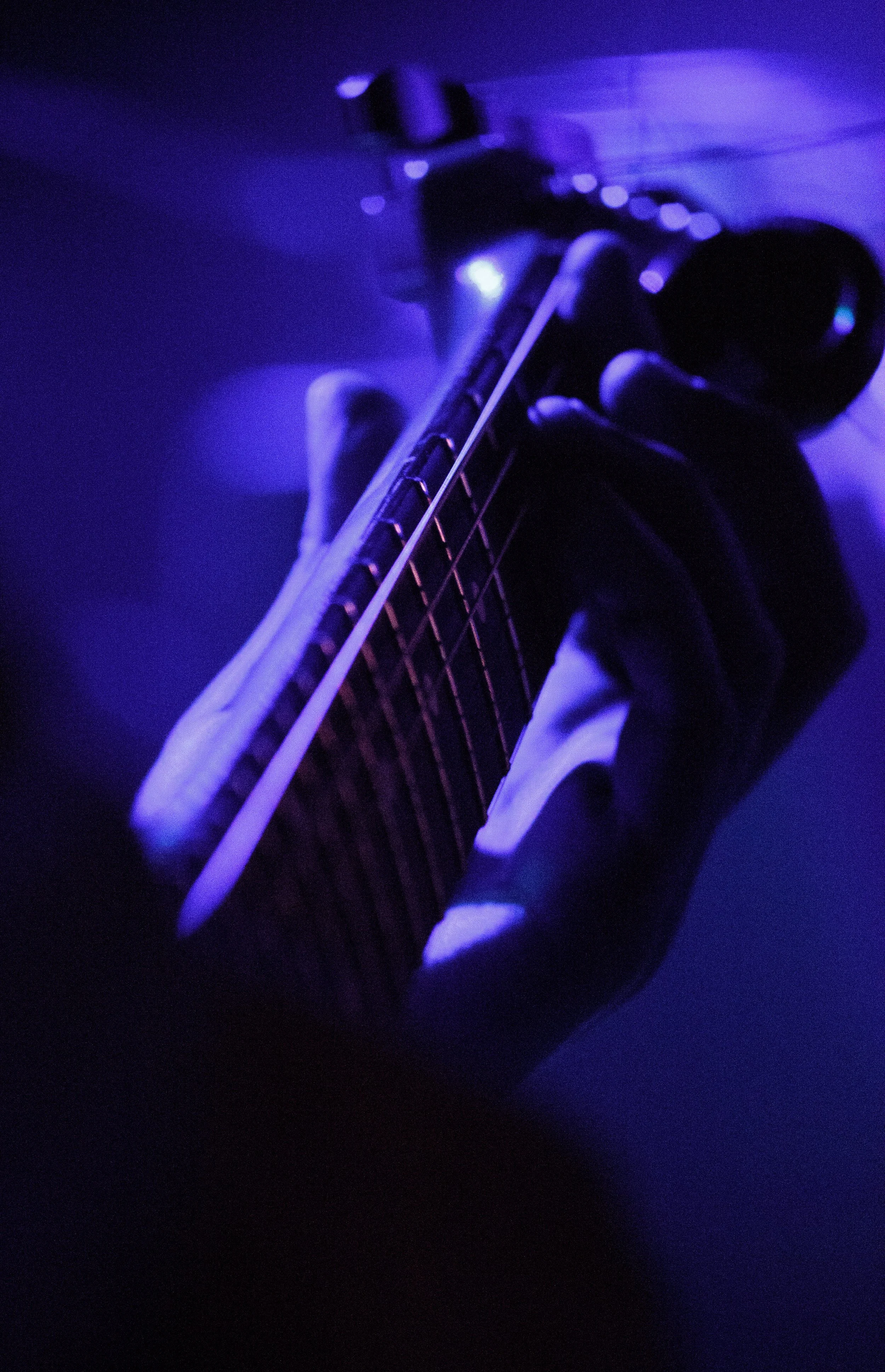 Close-up of a hand holding a guitar neck in a dark room illuminated by purple-blue light.