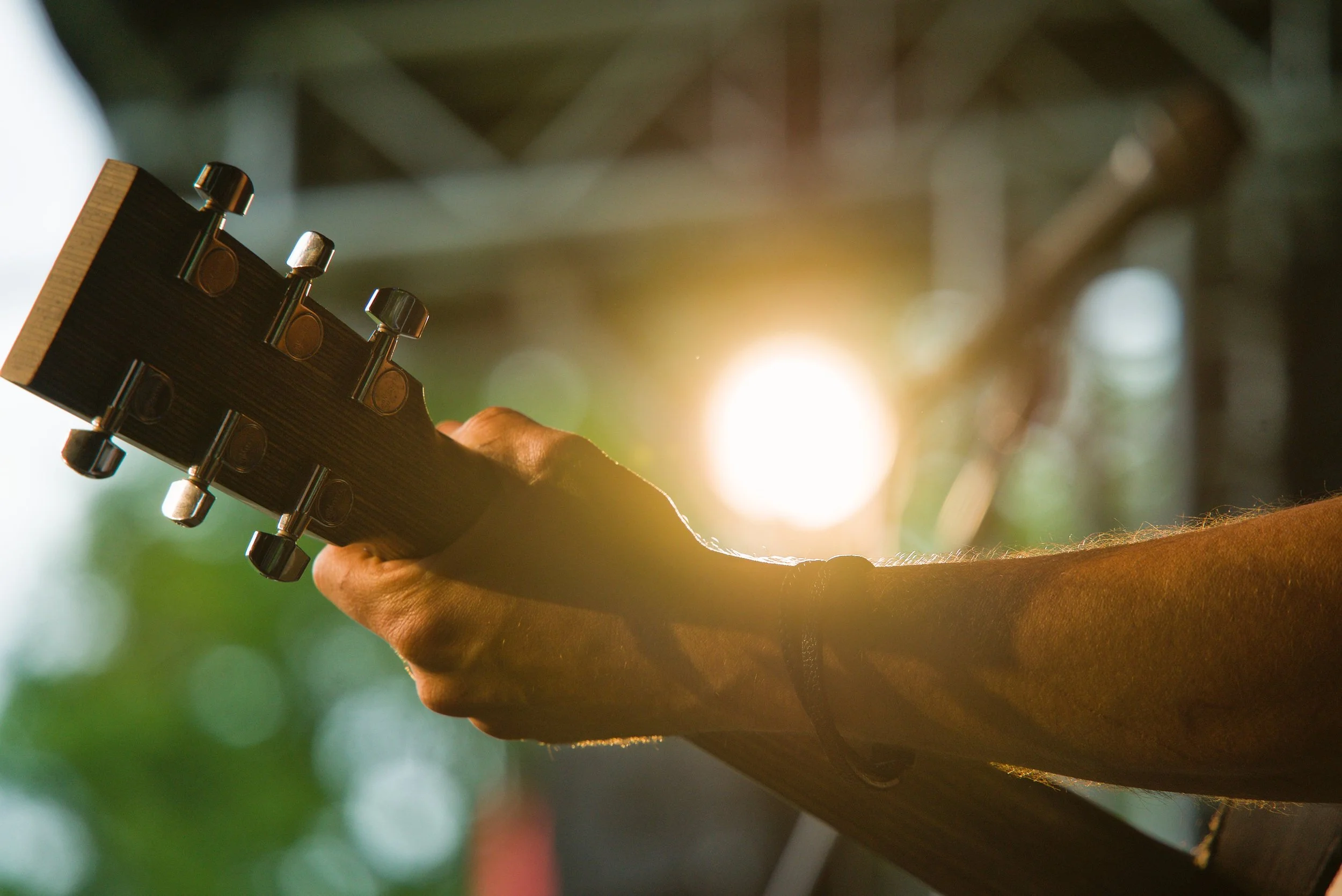 Person holding a guitar neck with tuning pegs, sunlight shining in the background, outdoor setting