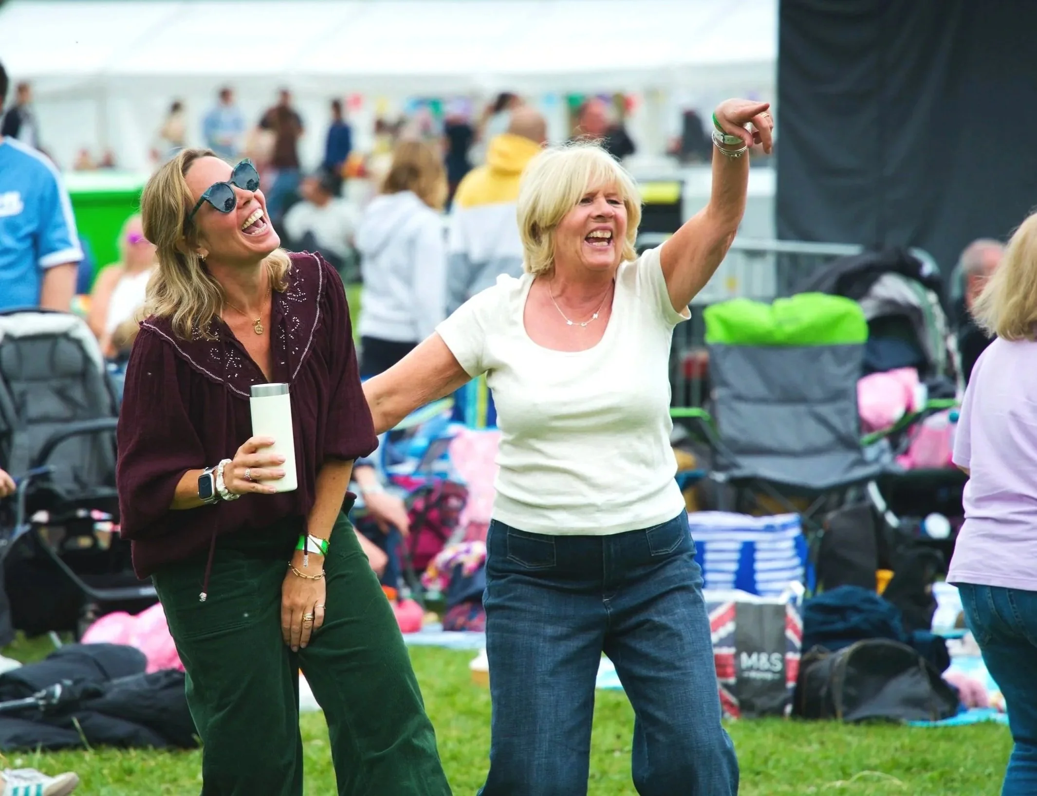 Two women smiling, dancing, and enjoying themselves at an outdoor event with tents and people in the background.