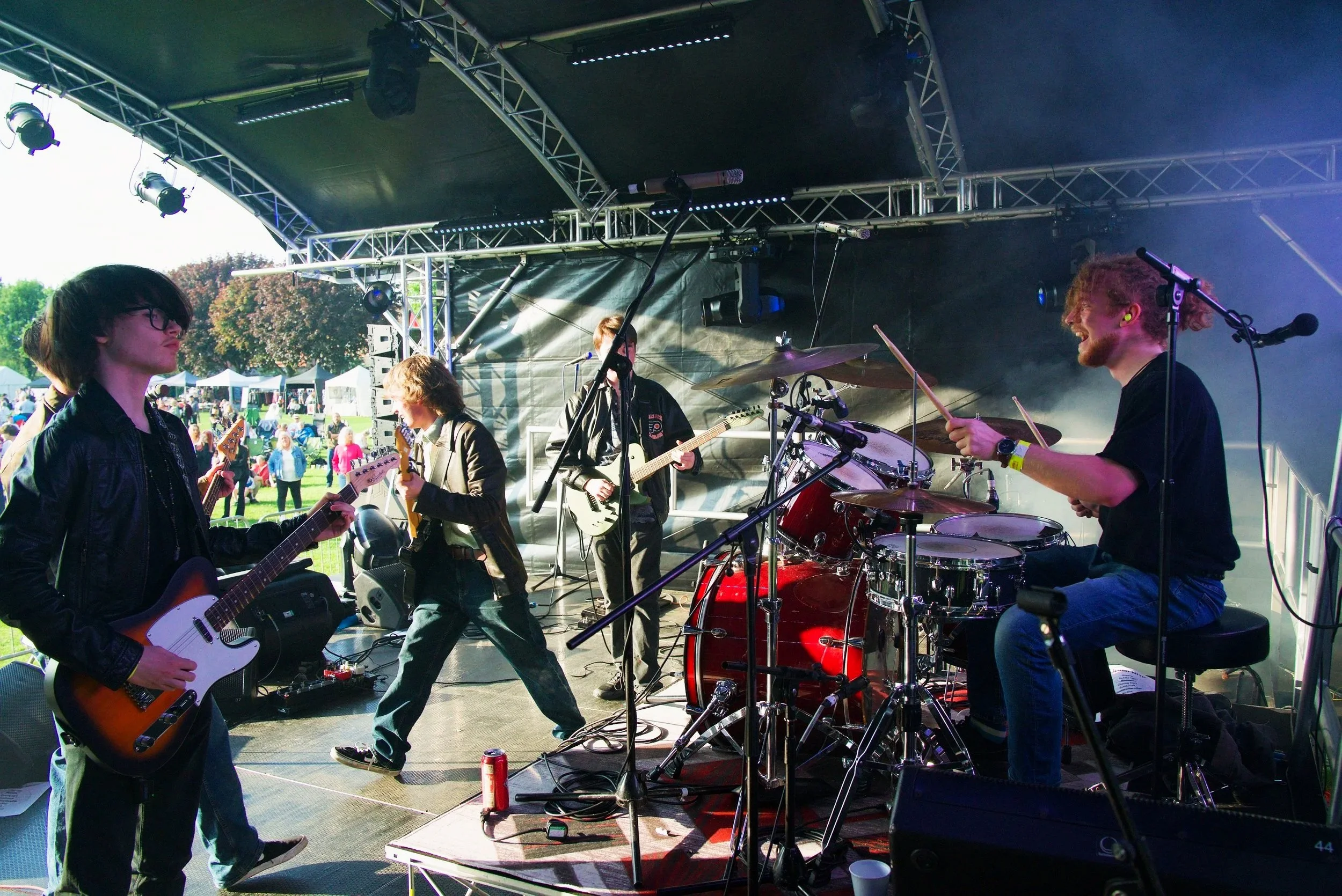 A band performing live on an outdoor stage with a drummer, two guitarists, and a bassist, surrounded by equipment and a crowd in the background.