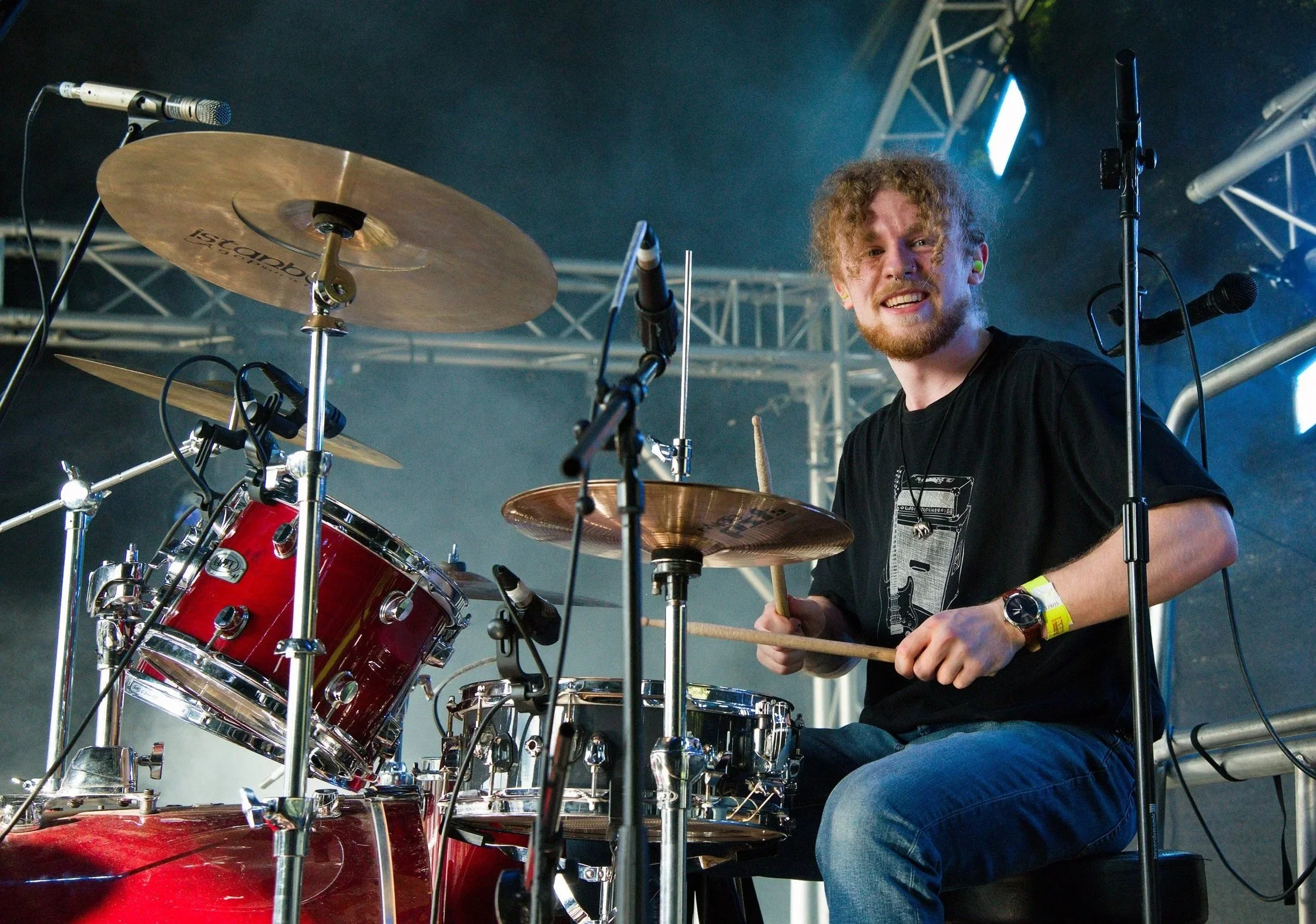 A male drummer with curly blonde hair and a beard, wearing a black t-shirt, playing a red drum set on stage surrounded by microphones and stage lighting.