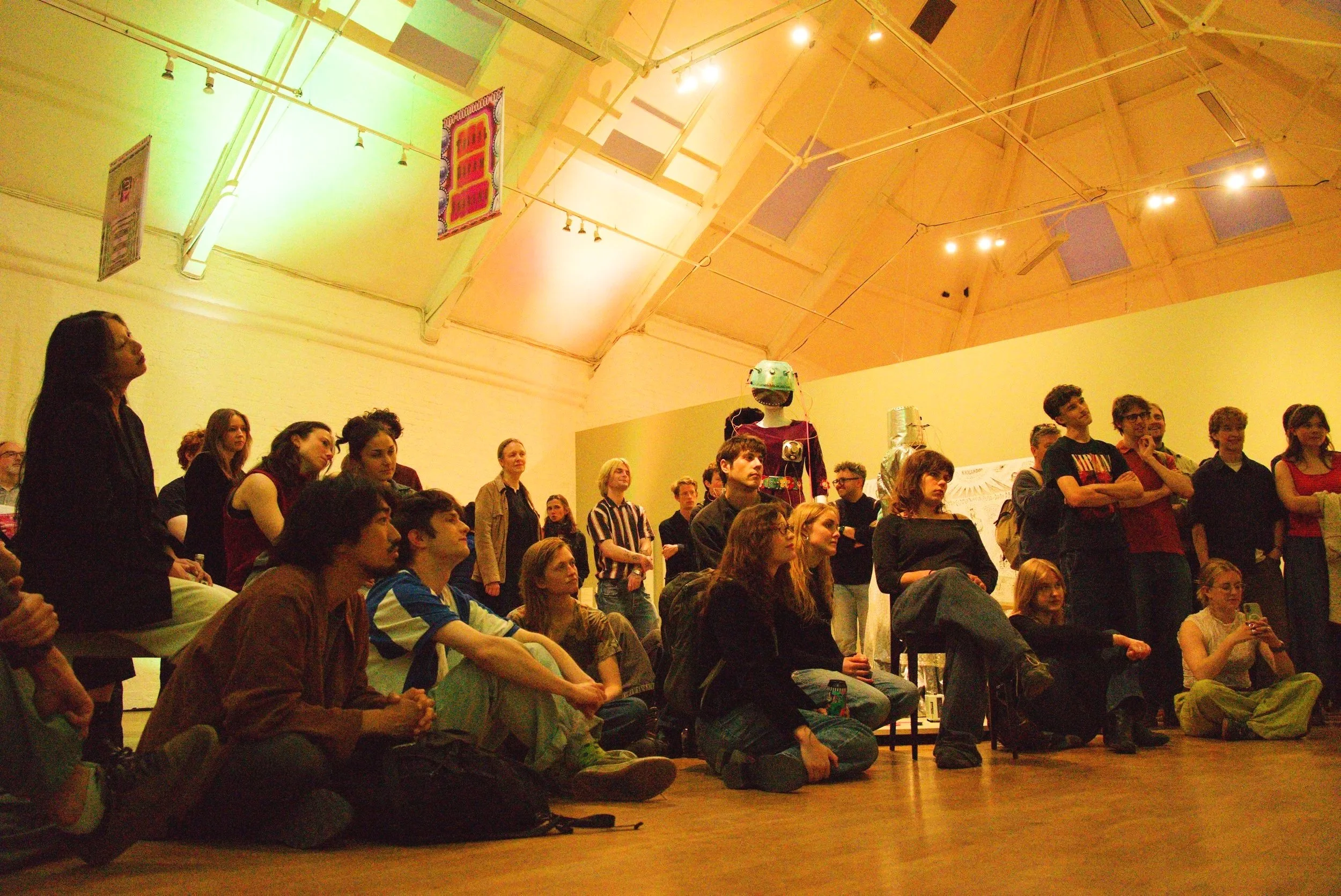 A group of people sitting and standing in an art gallery or museum, watching an exhibit or presentation. The diverse crowd appears attentive, with some sitting on chairs and others standing at the back. The ceiling has colorful lighting and banners, 