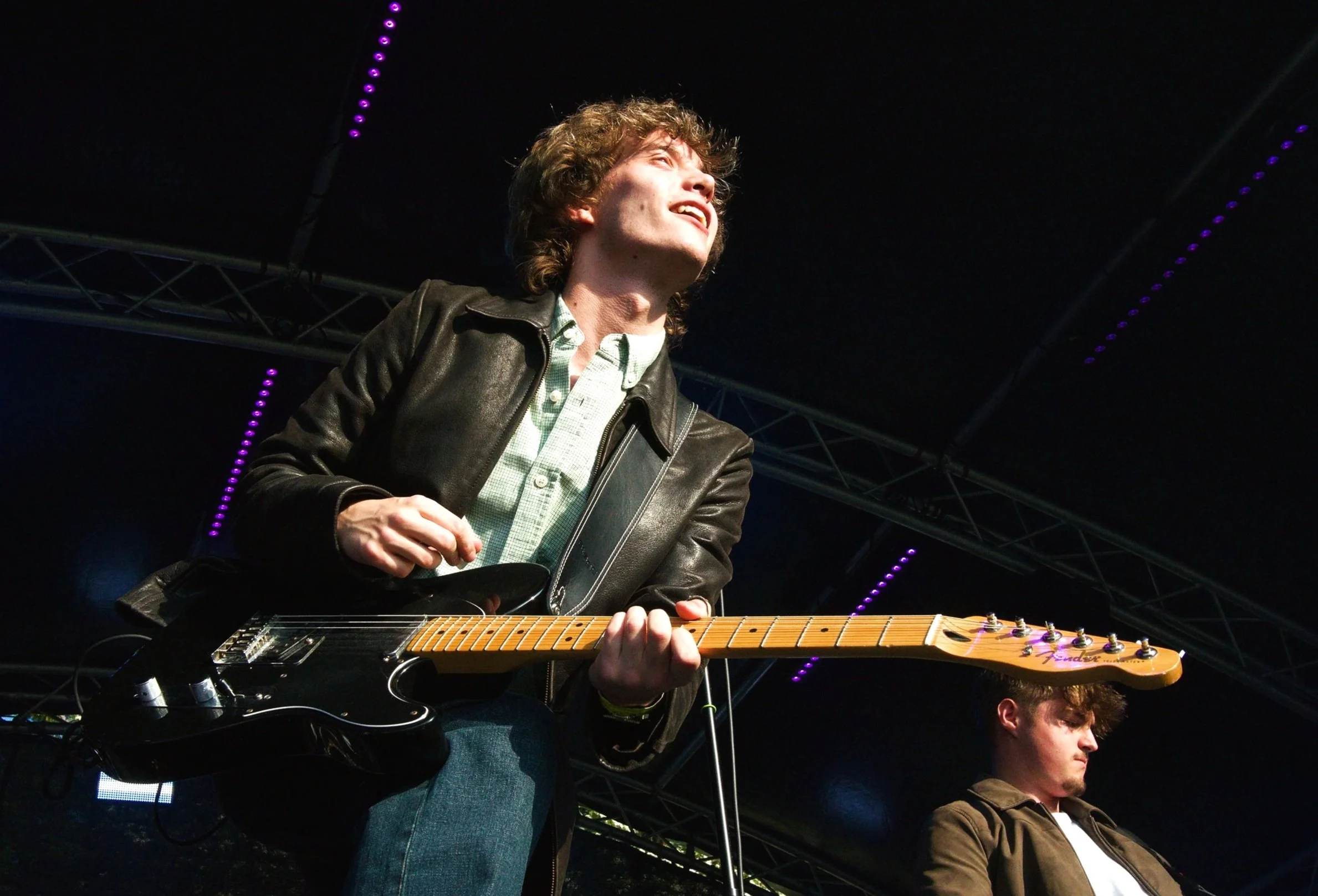 A person playing an electric guitar on stage, illuminated by stage lights, with another person in the background.