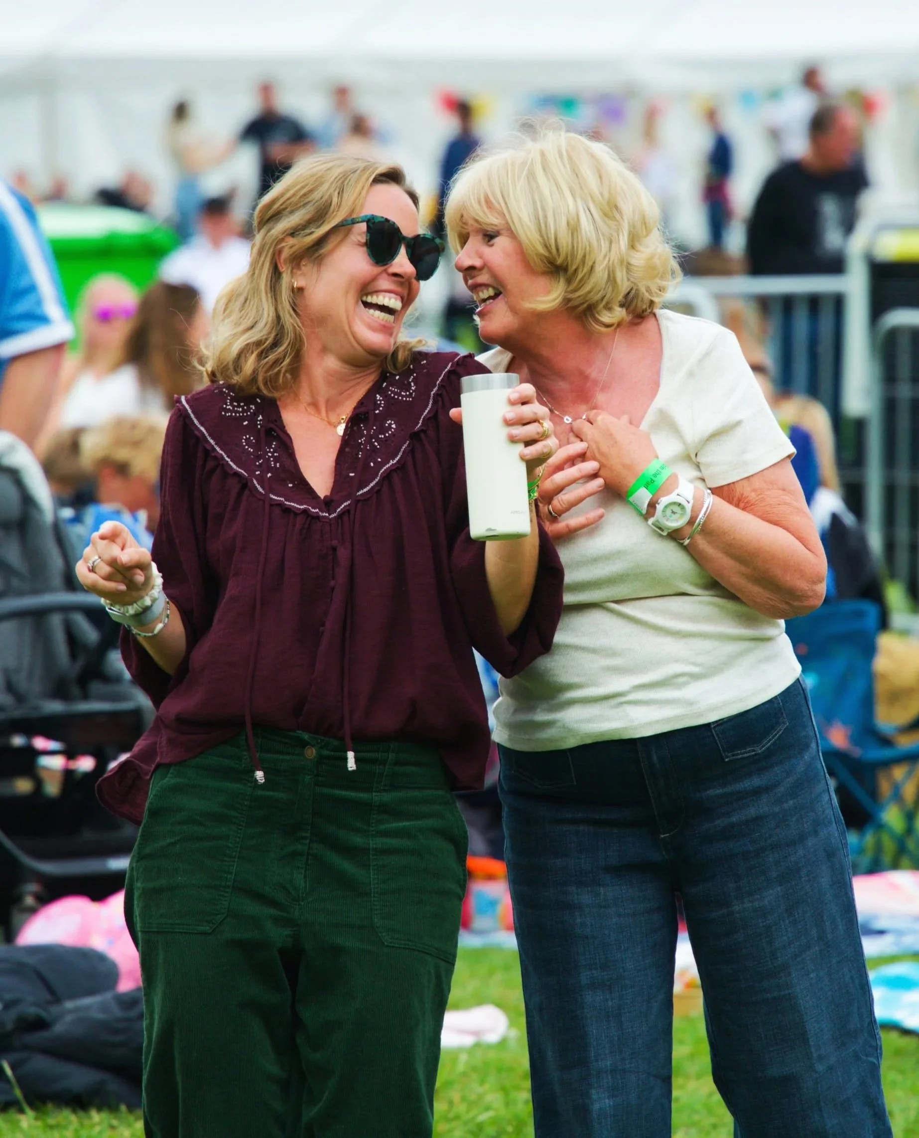 Two women laughing and talking at an outdoor event, with a crowd and tents in the background.