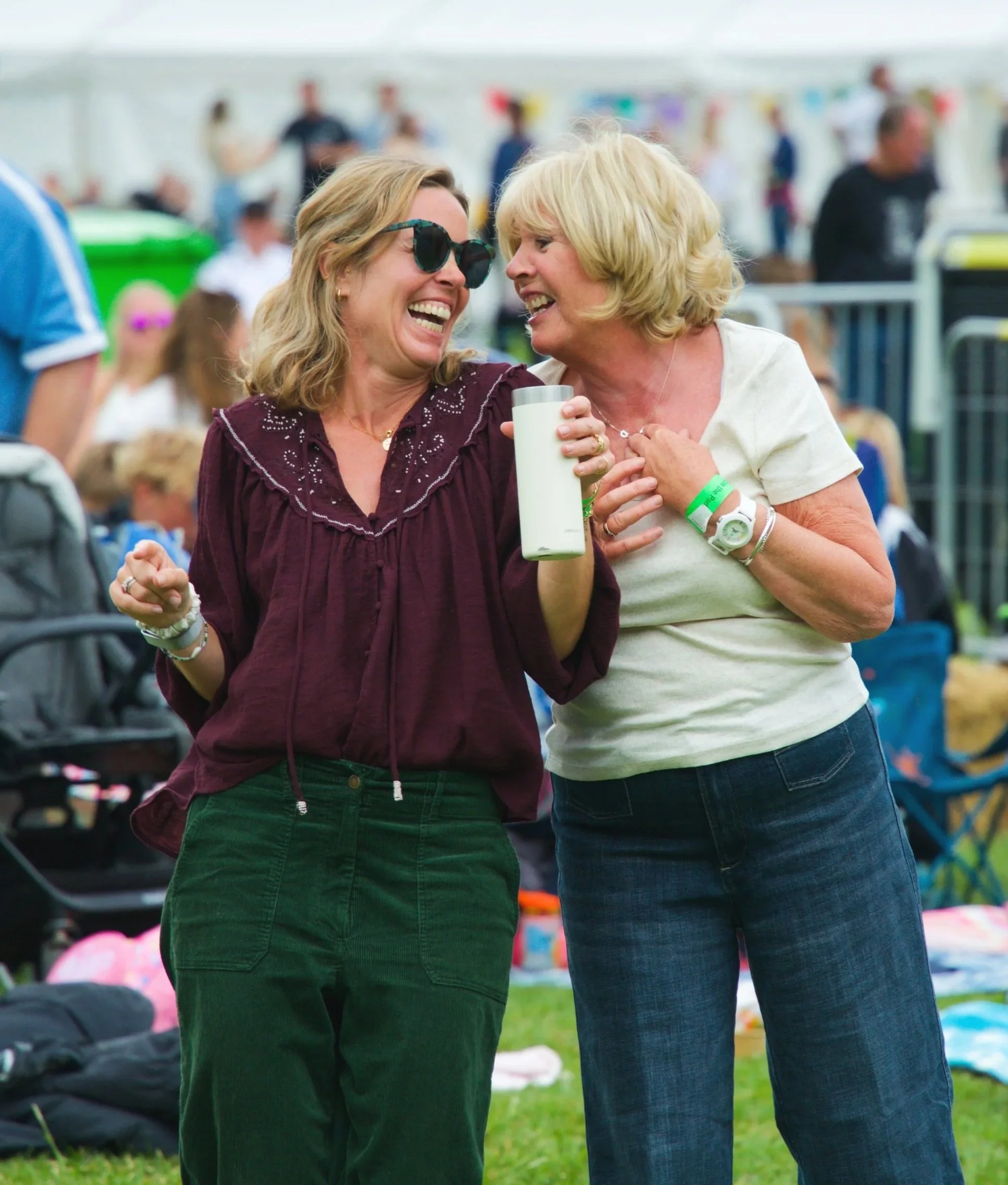 Two women sharing a joyful moment at an outdoor event, smiling and laughing while holding a drink, with a blurred crowd and colorful decorations in the background.