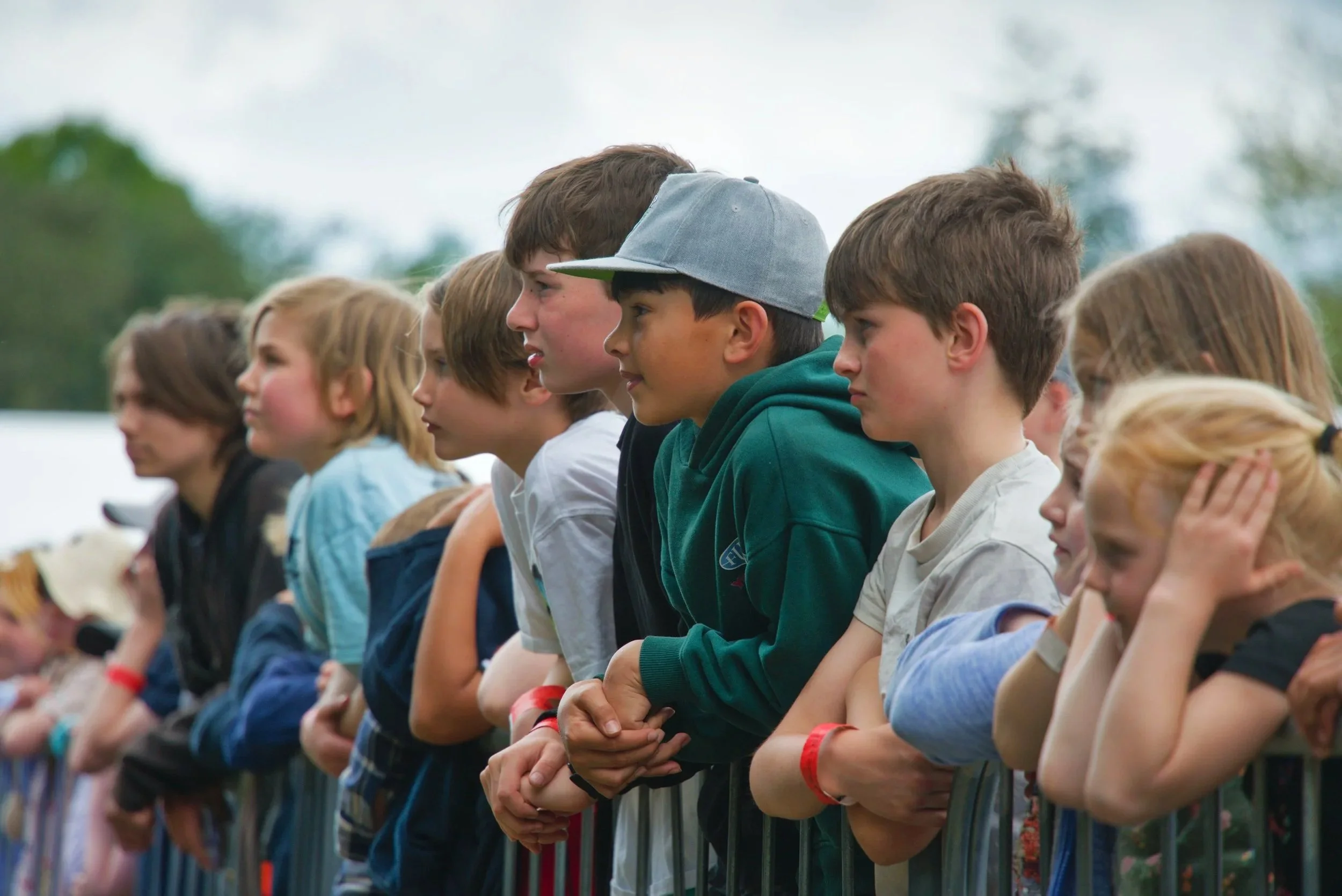 A group of children standing side by side outdoors, leaning on a metal fence, some with eyes closed or hands clasped, appearing to be attending an event or watching something.