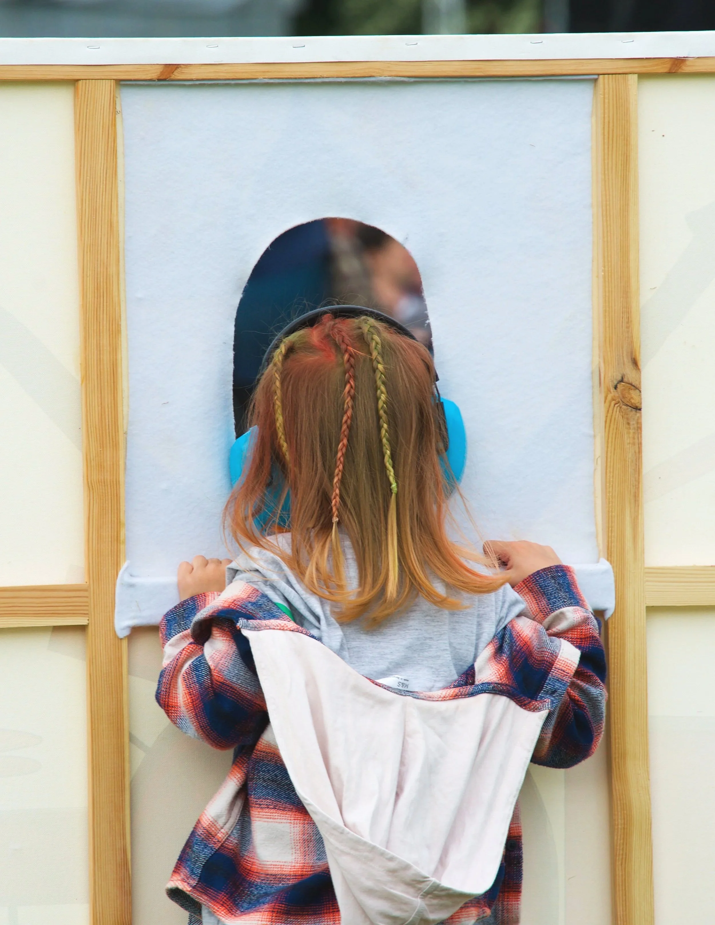 A young girl with braided hair peeks through a hole in a cardboard box house.