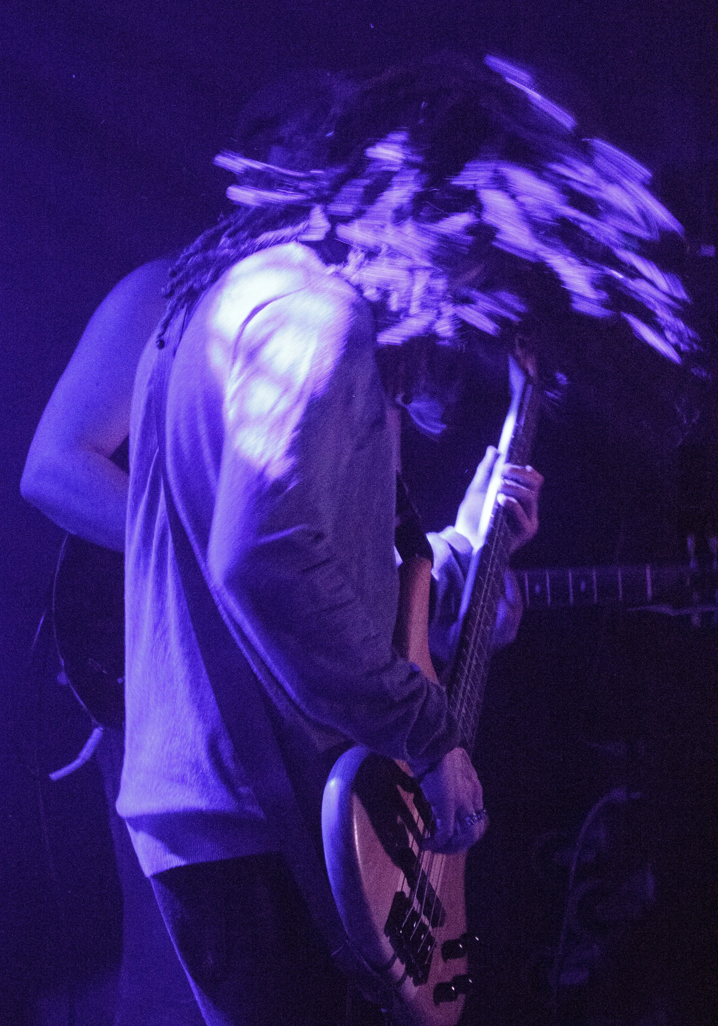 Person with long, bushy hair playing an electric guitar in a dimly lit space illuminated with purple light.