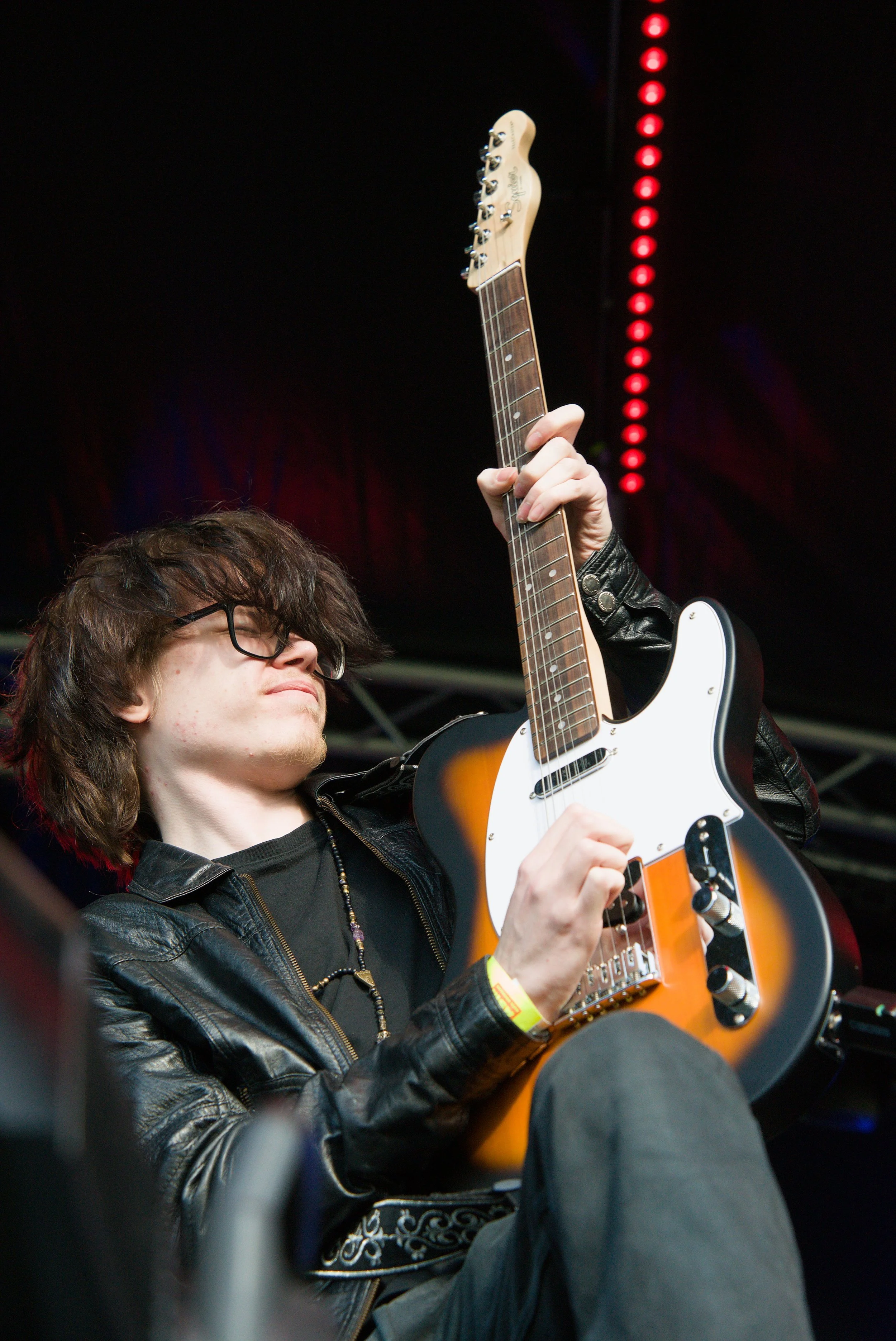 A young person with long brown hair, glasses, wearing a black leather jacket, playing an orange and white electric guitar on stage during a performance.
