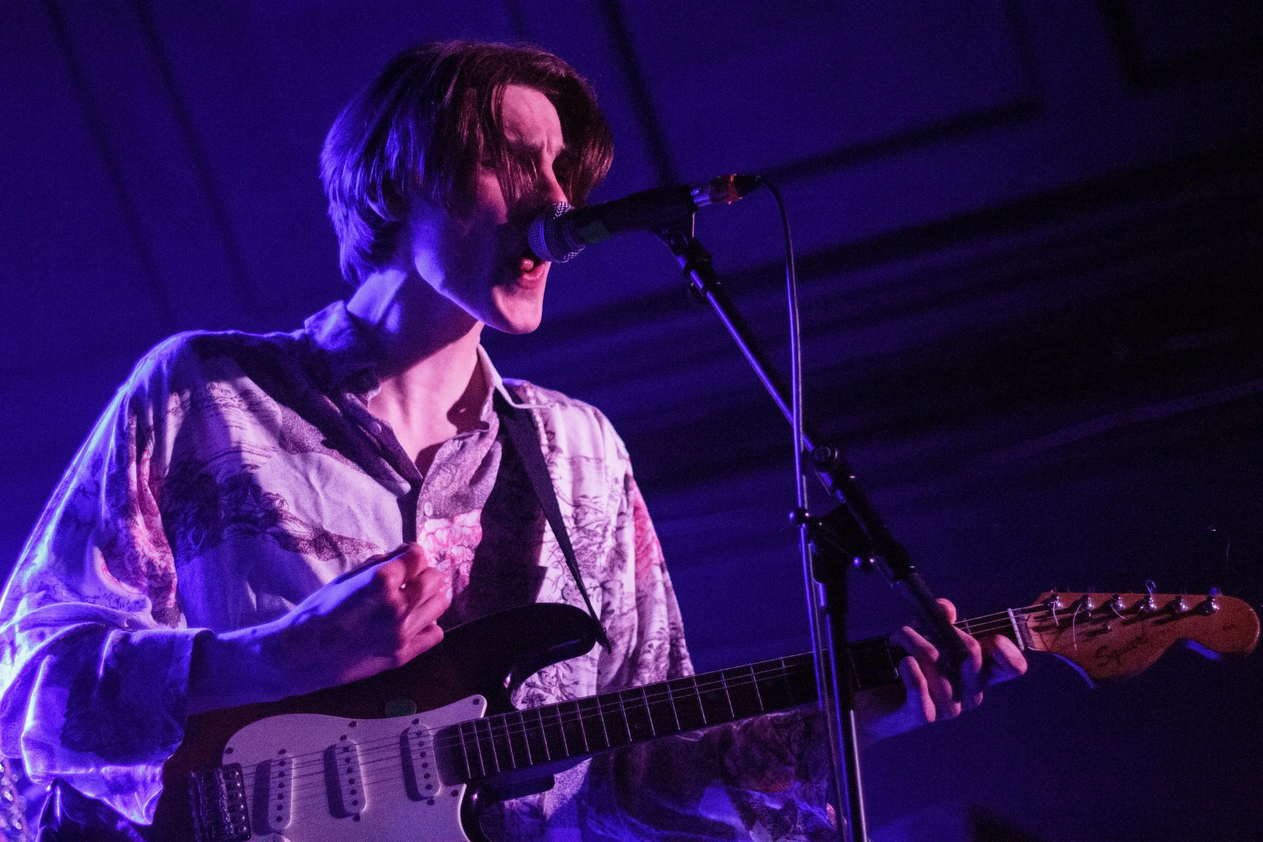 A person with shoulder-length hair singing into a microphone while playing an electric guitar on stage, illuminated by purple stage lights.