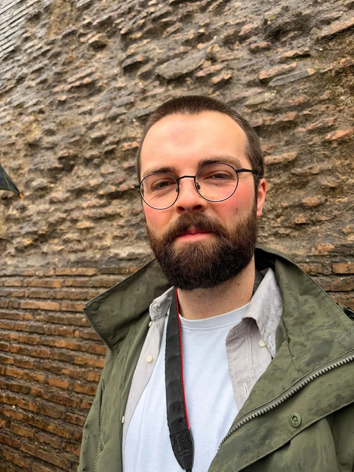 A bearded man with glasses taking a selfie in front of a textured brick wall. He is wearing a light-colored shirt and a green jacket, with a camera strap around his neck.