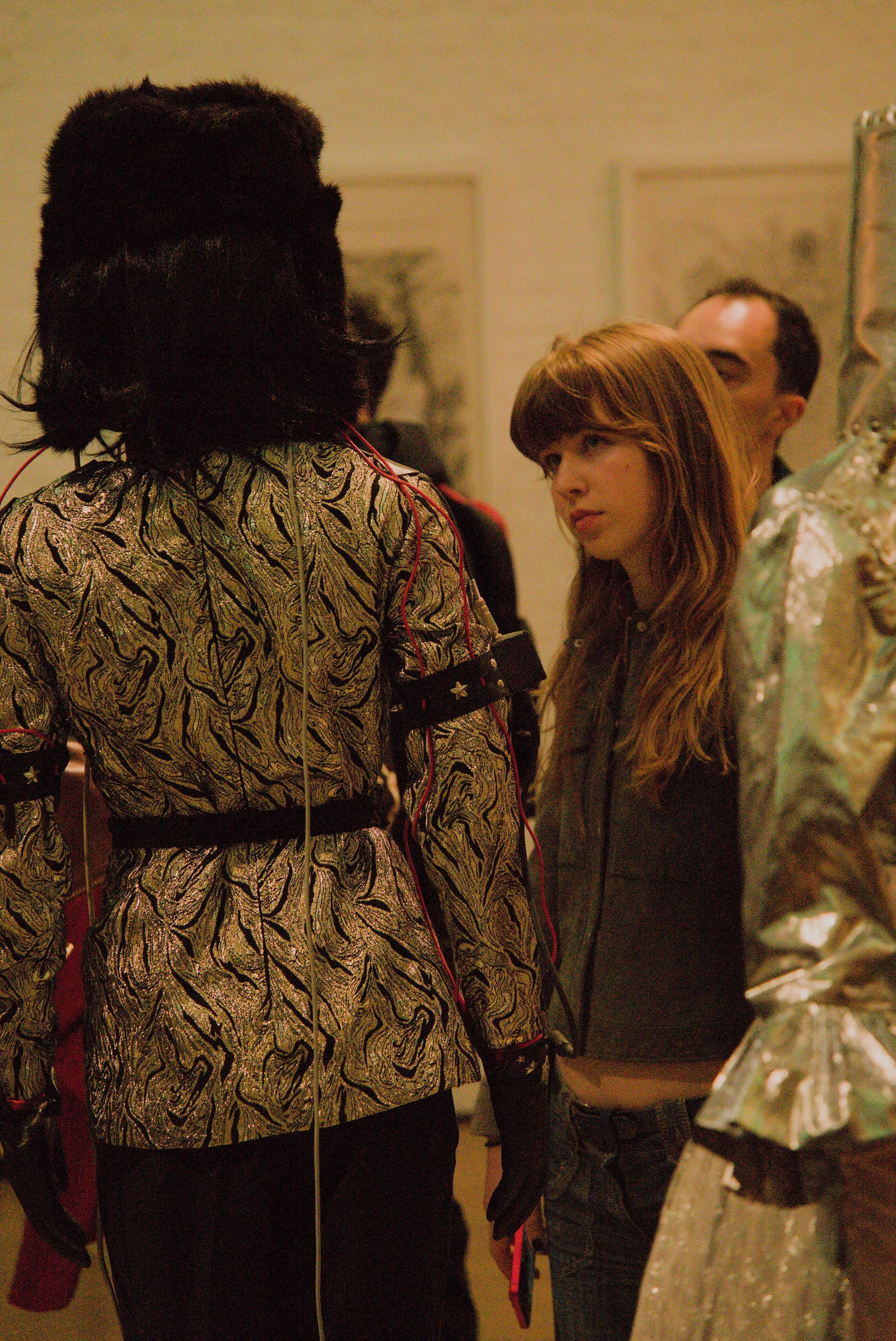 A group of people are standing and talking in an indoor setting, with varied clothing styles and hair colors, including black, brown, and red hair.