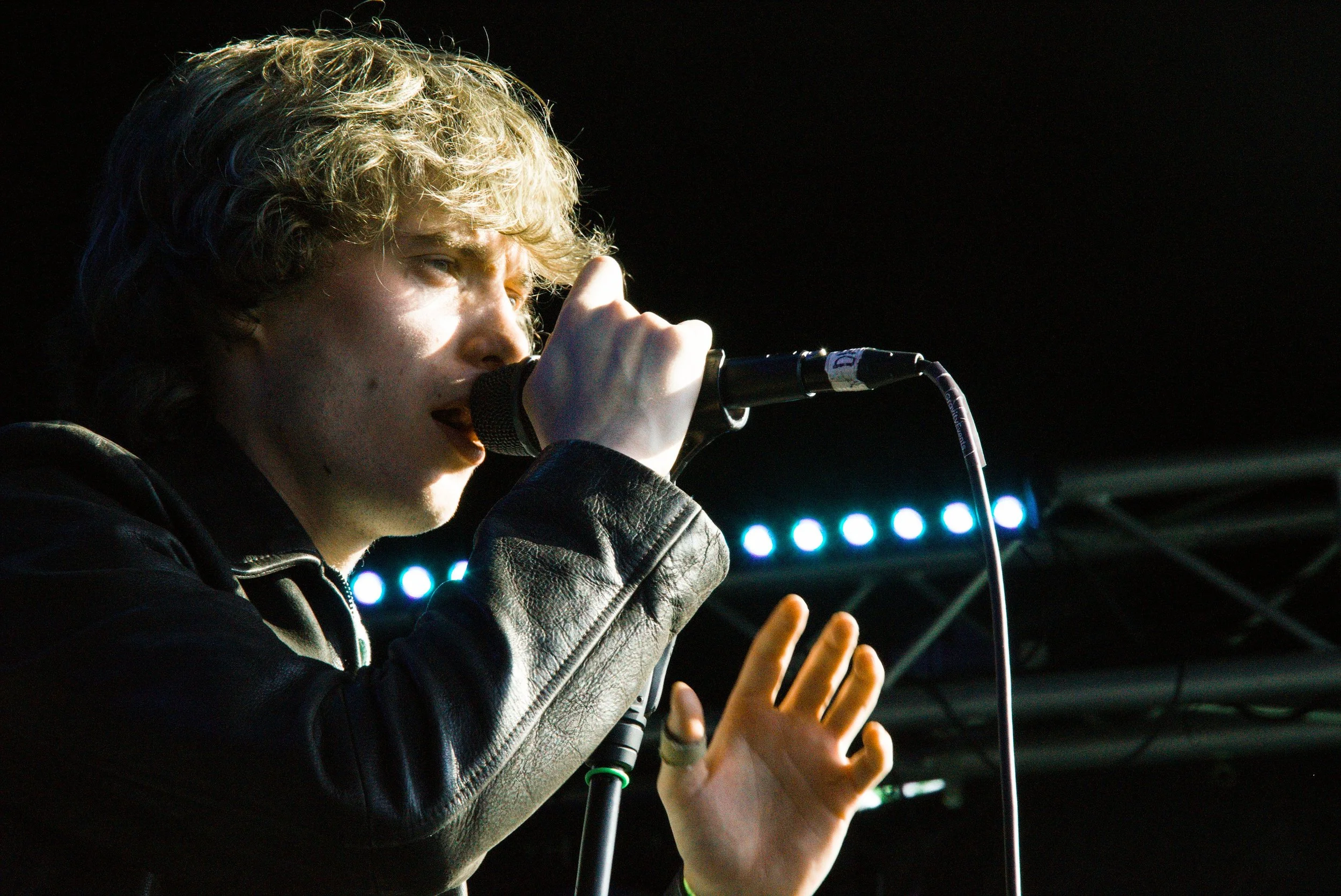 A young man singing into a microphone on stage, with his hands raised, wearing a black leather jacket and a ring on his finger.