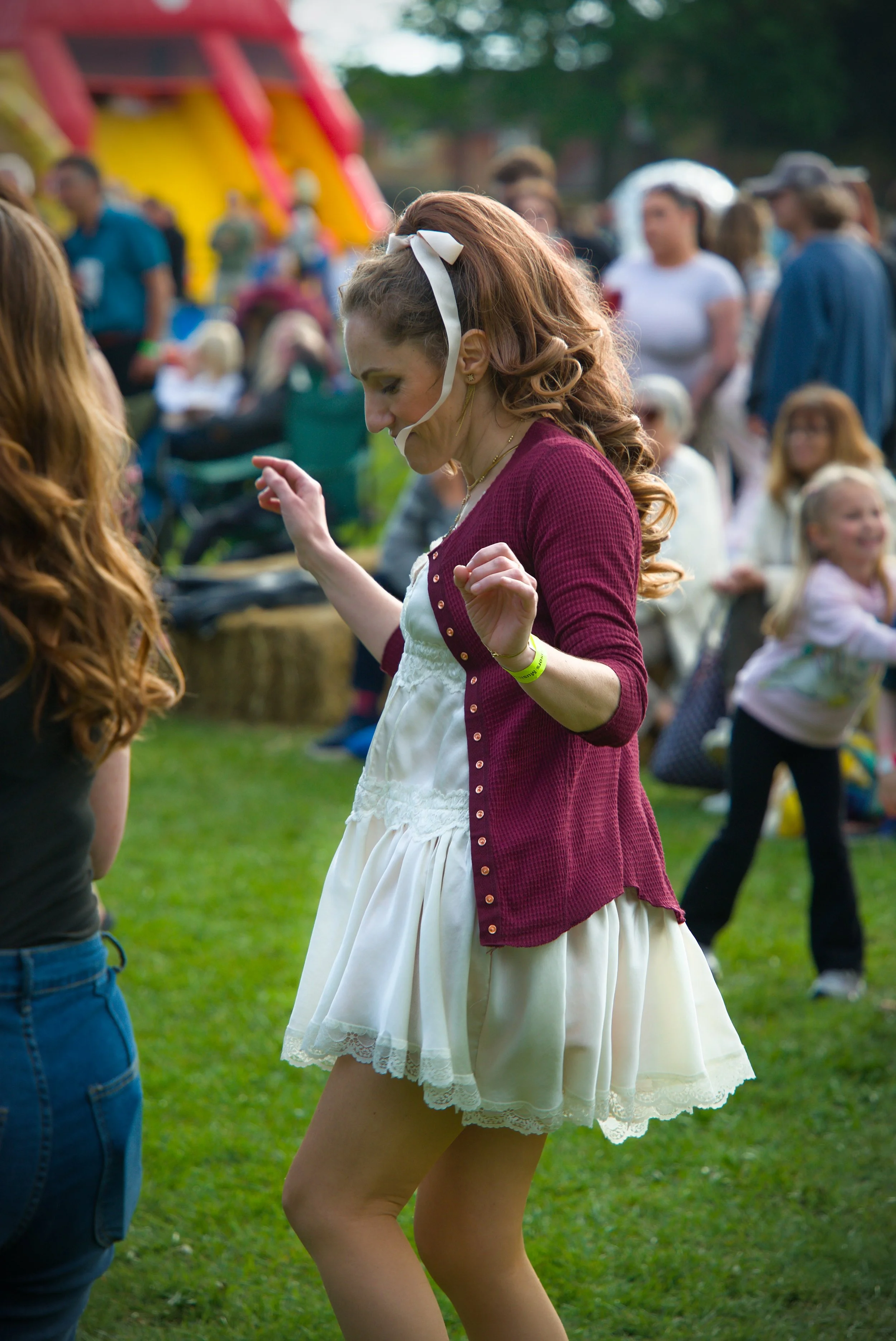 A young woman with curly red hair, wearing a white dress with lace and a red cardigan, dancing outdoors at a festival or fair.