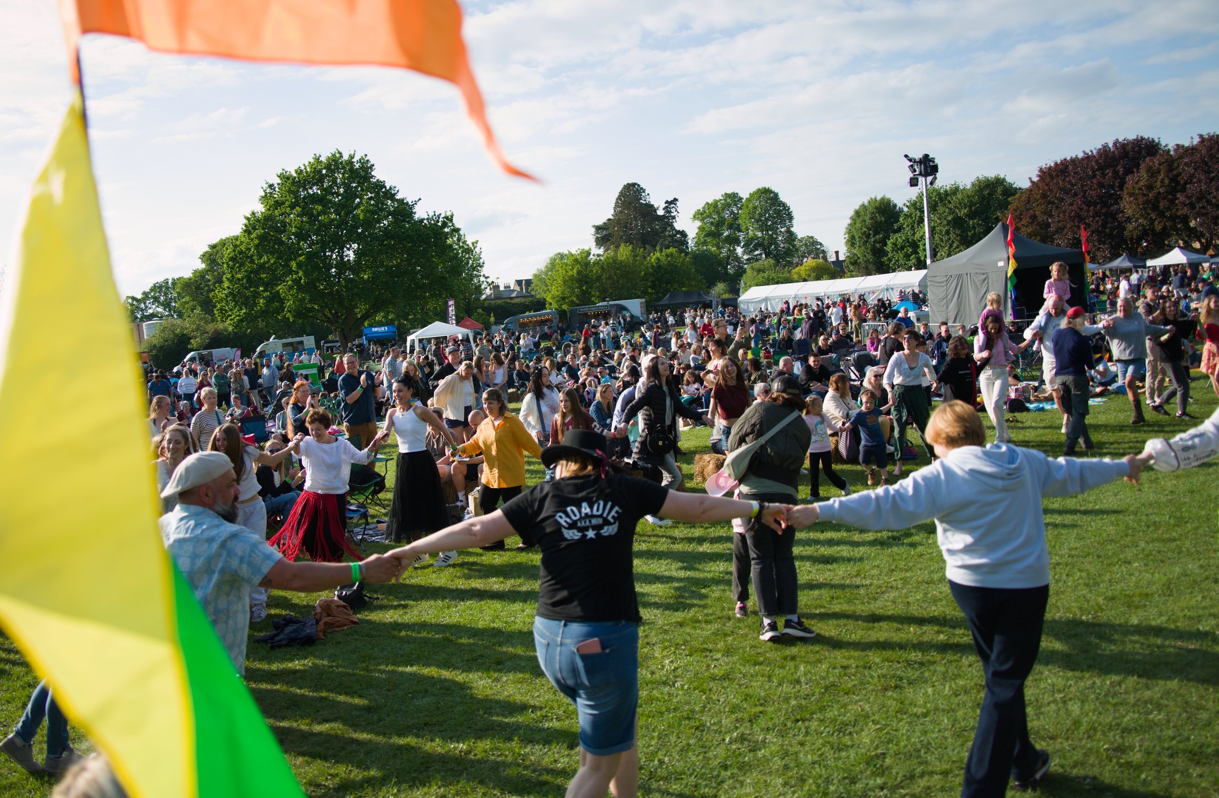 People holding hands and dancing in a large outdoor park during a festival or celebration, with tents, trees, and a blue sky in the background.