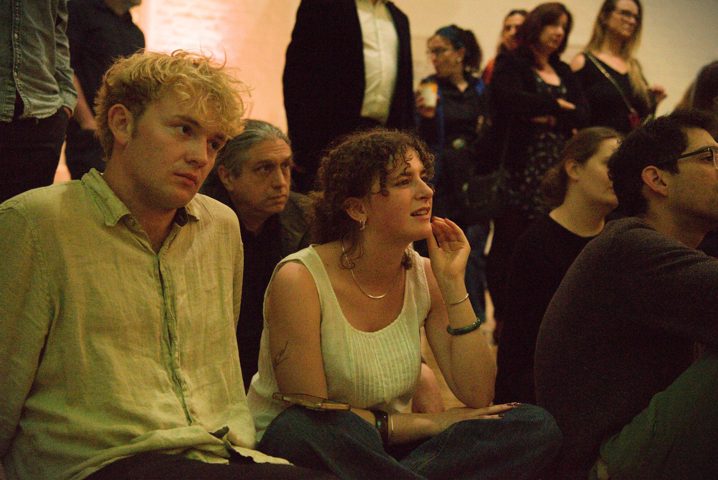 People seated and standing, attending an indoor event, with diverse hairstyles and clothing, engaged and listening.