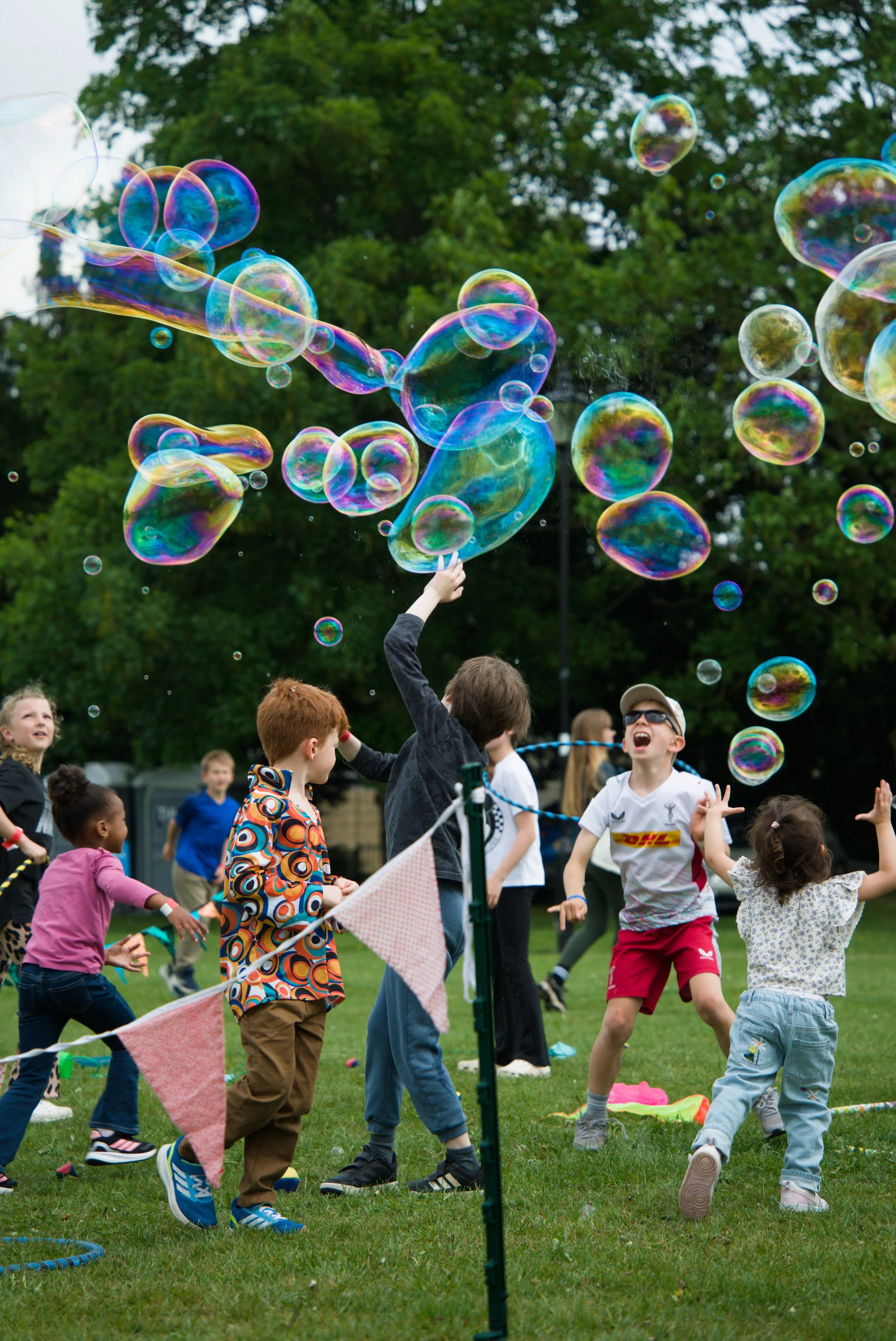 Children playing and laughing outdoors during a bubble-blowing activity on a grassy field with trees in the background.